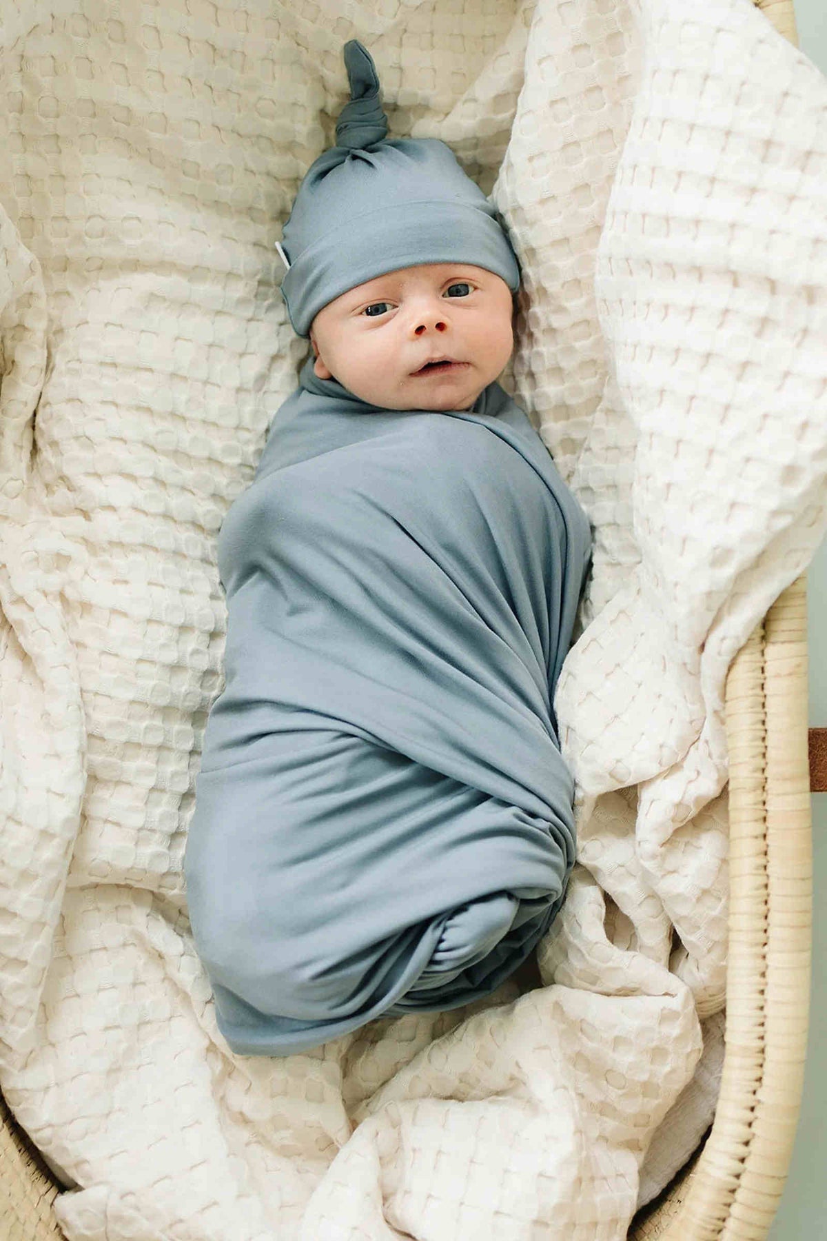 A baby wrapped in a dusty blue swaddle blanket with a matching hat lies on a textured white blanket.