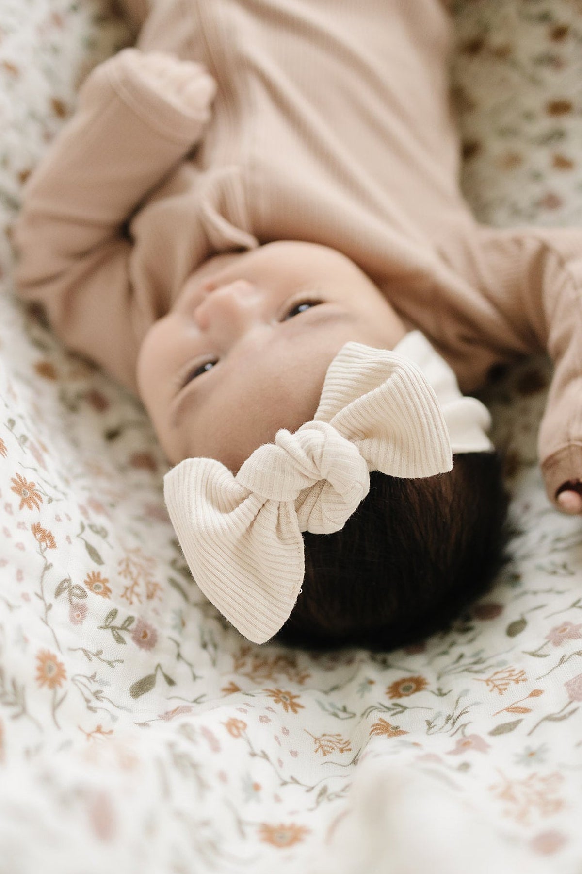 A baby wearing a vanilla ribbed head wrap lies on a floral-patterned blanket, dressed in a beige outfit.