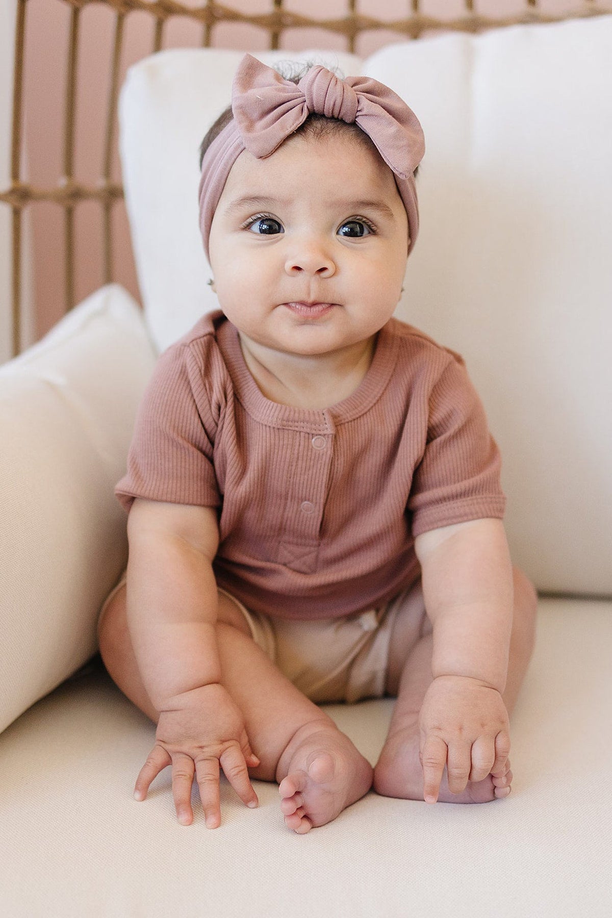A baby wearing a dusty rose head wrap and matching top sits on a white cushioned chair.