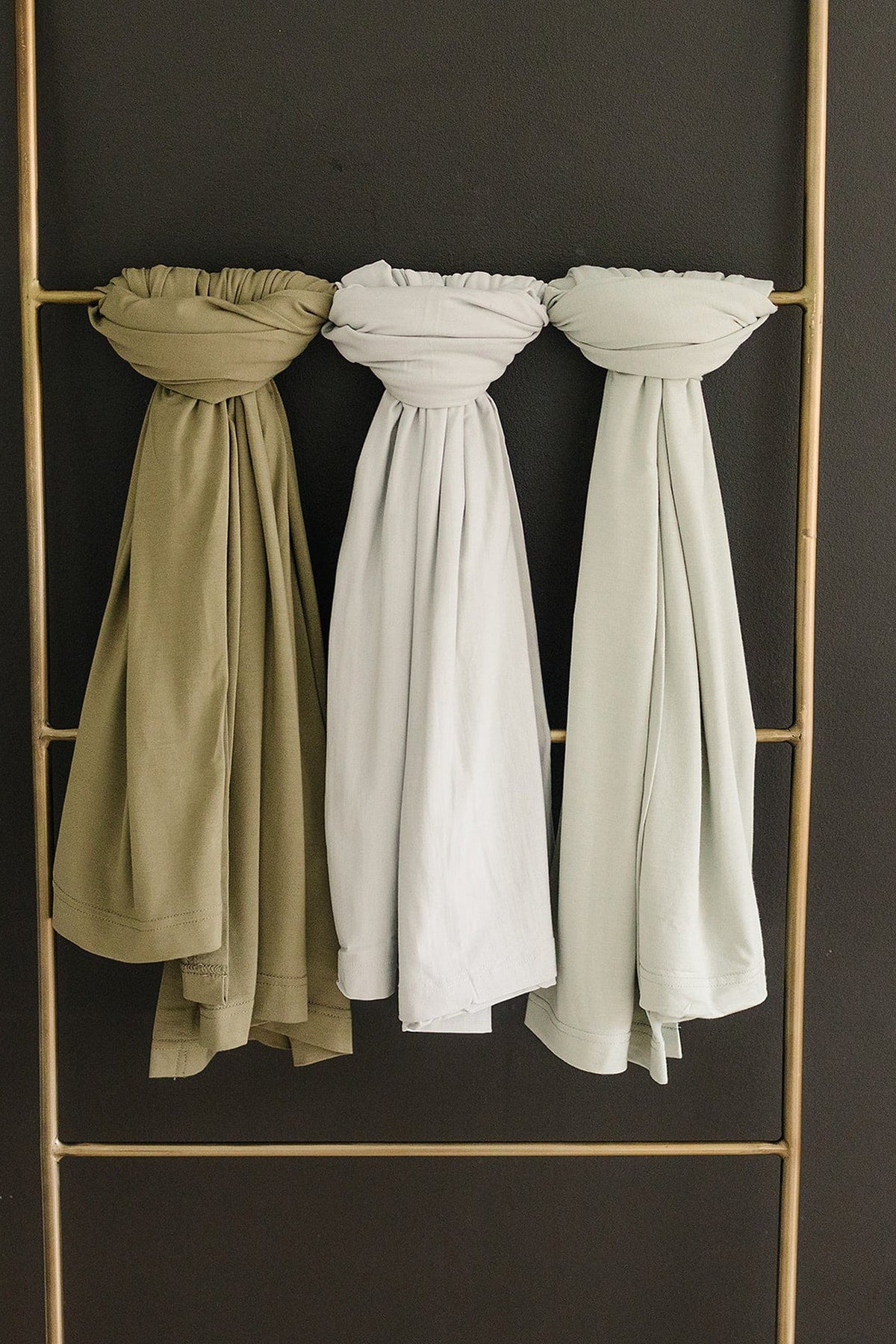 Three bamboo swaddle blankets in olive, white, and light sage hang on a brass rail against a dark background.