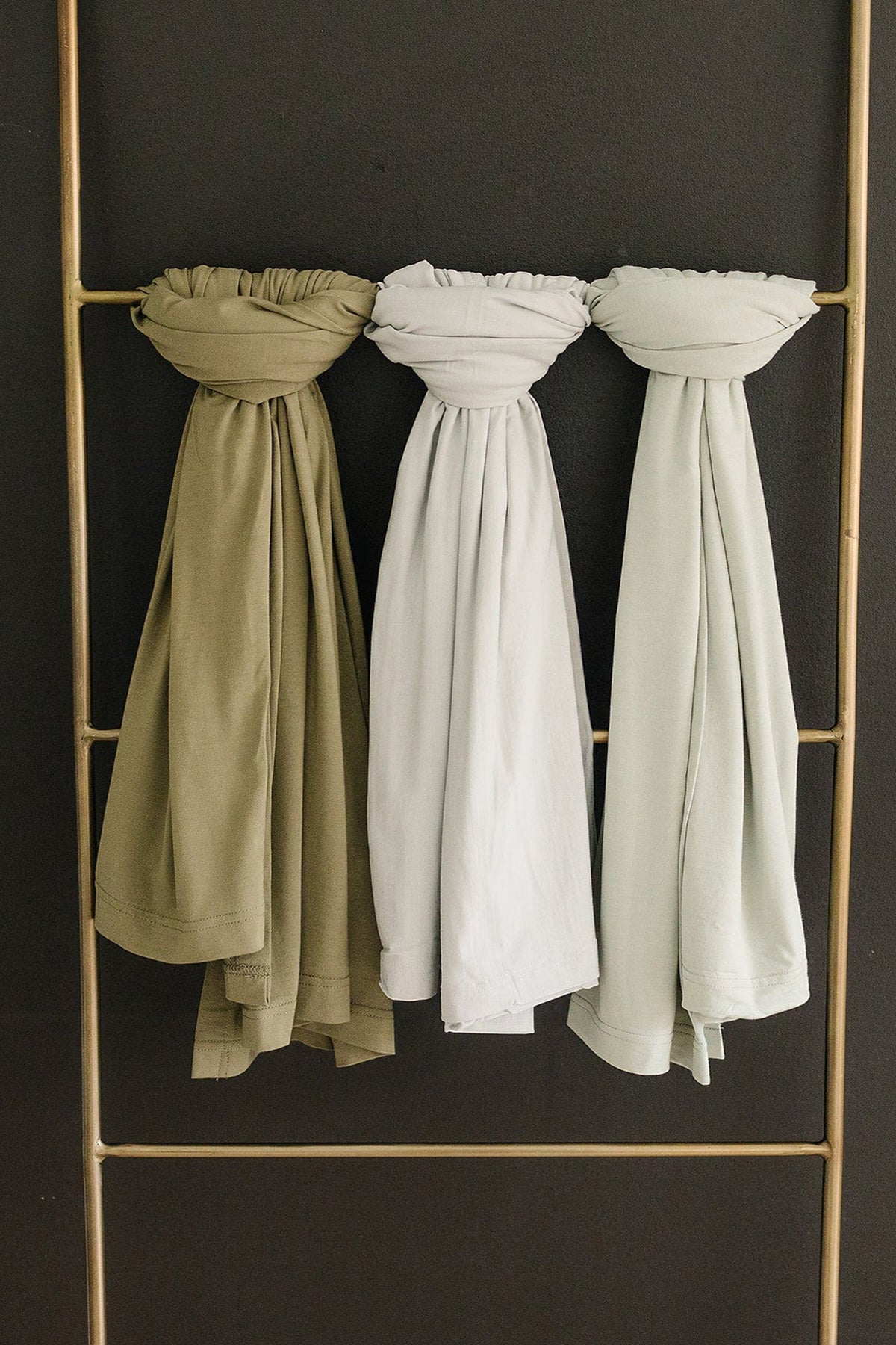 Three swaddle blankets in sage, white, and light green hang neatly on a brass rod against a dark wall.