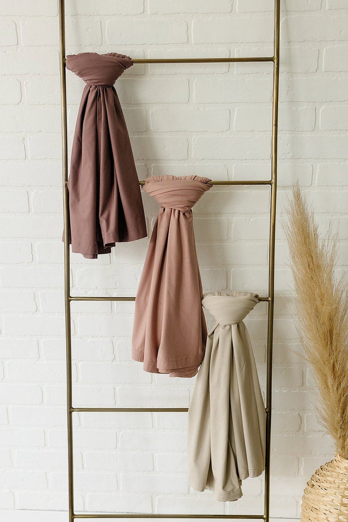 Three swaddle blankets in brown, dusty rose, and beige hang on a metal ladder against a white brick wall.