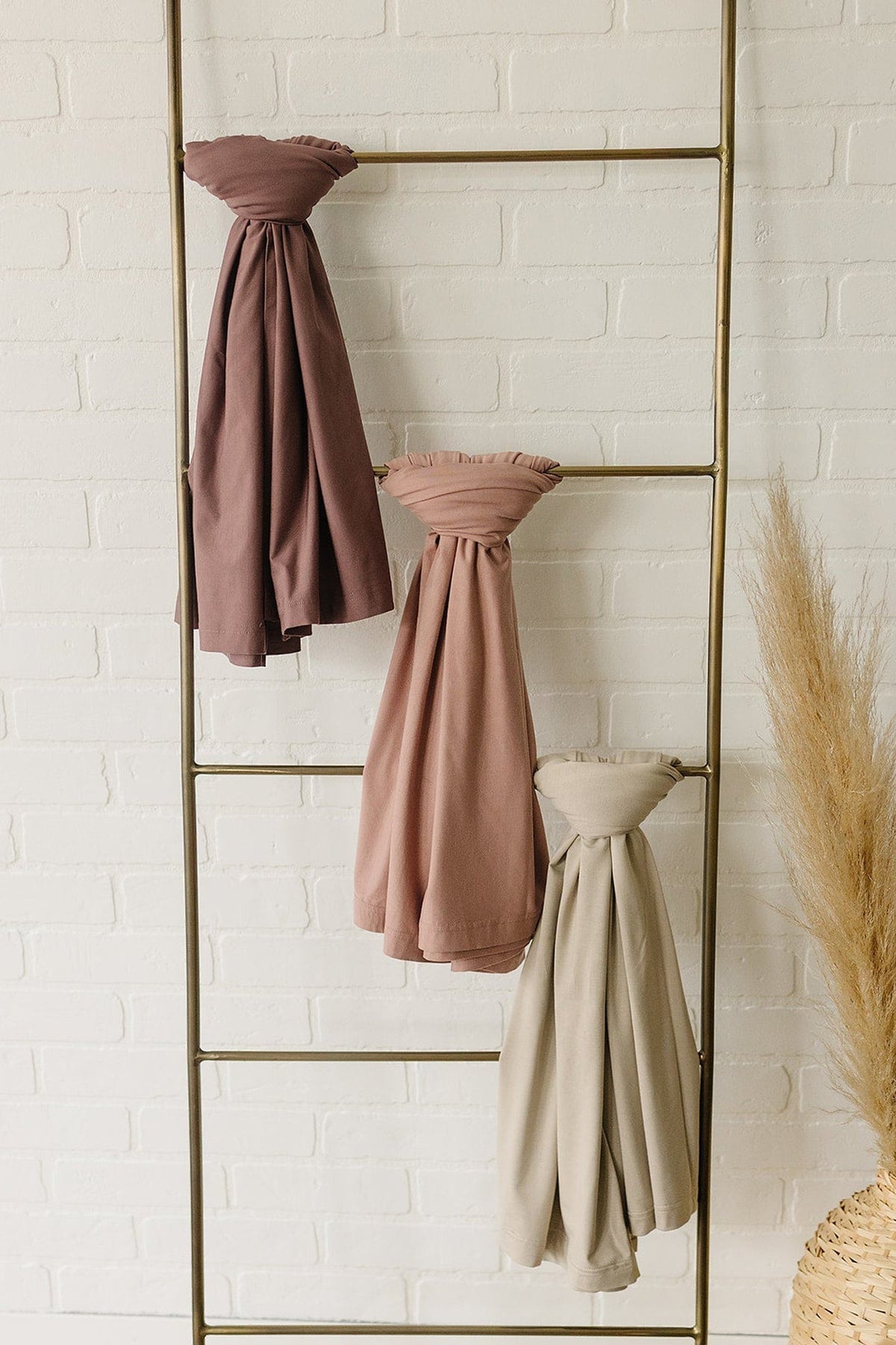 Three bamboo swaddle blankets in mauve, pink, and beige hang on a brass ladder against a white brick wall.