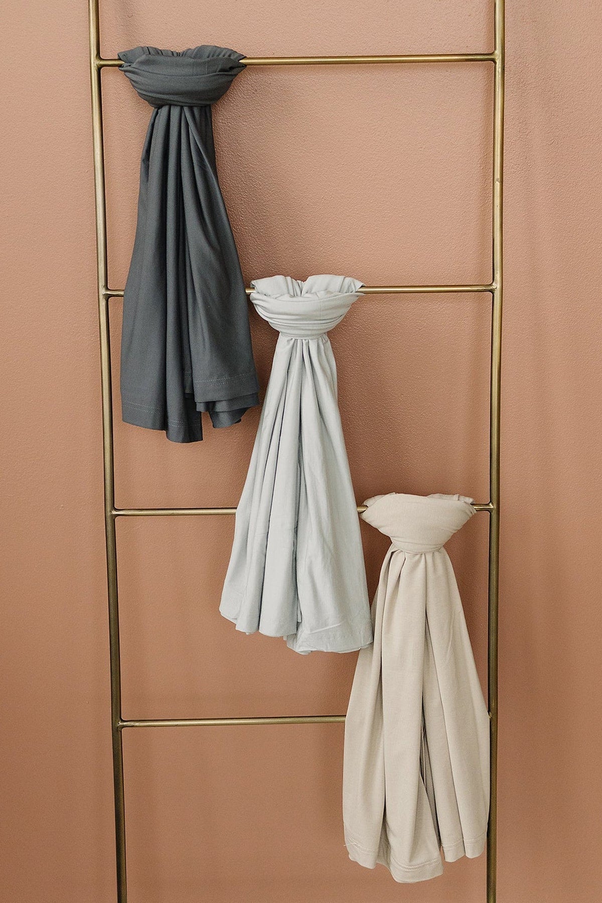 Three swaddle blankets in charcoal, light gray, and beige hang on a brass ladder against a peach wall.