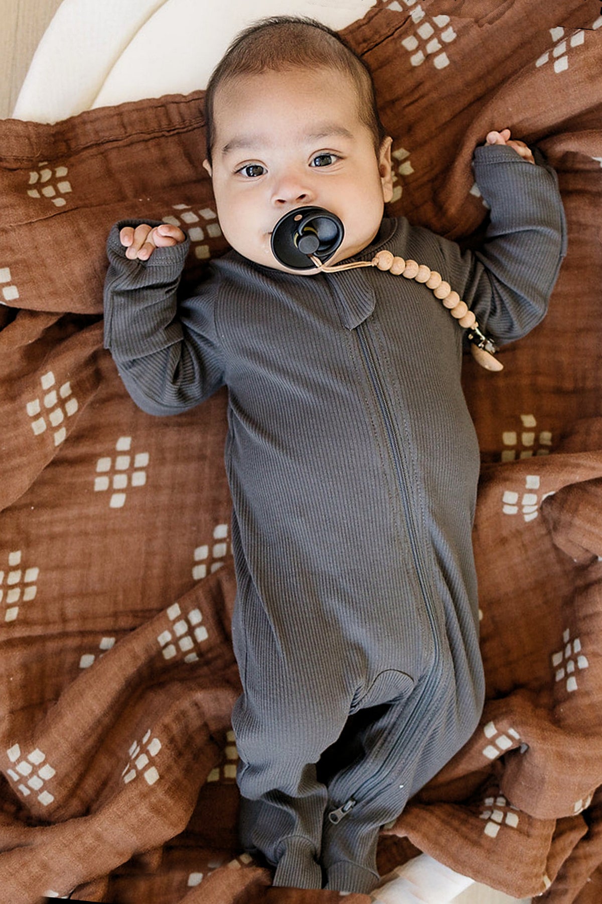 A baby lies on a patterned brown blanket, wearing a charcoal ribbed zipper footed pajama with a pacifier.
