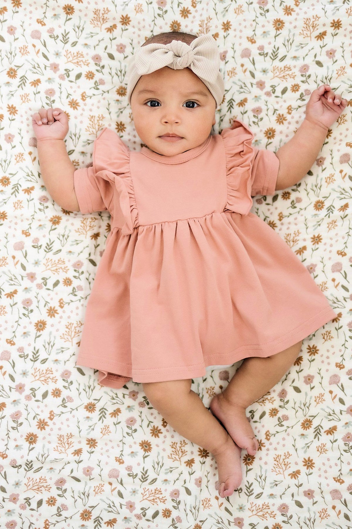 A baby in a pink dress and beige ribbed head wrap lies on a floral-patterned blanket.