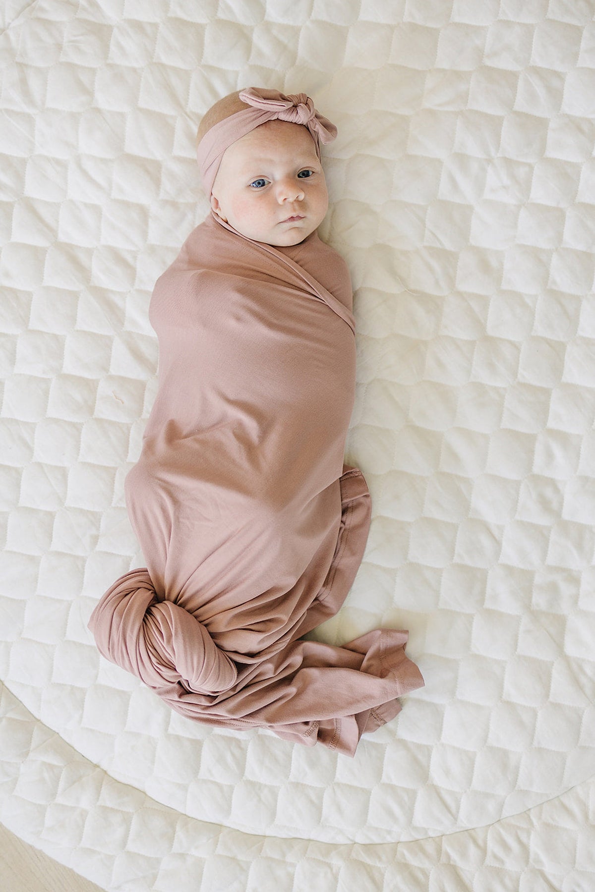 Baby wrapped in a dusty rose swaddle blanket with matching headband, lying on a quilted white blanket.