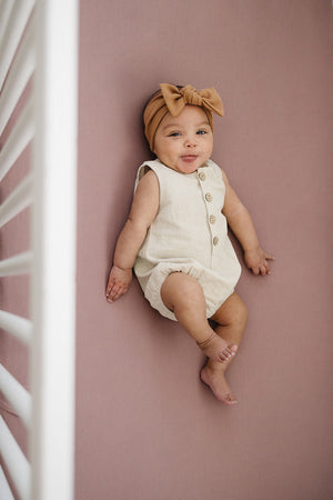 A baby in a beige romper and brown headband lies on a dusty rose crib sheet.