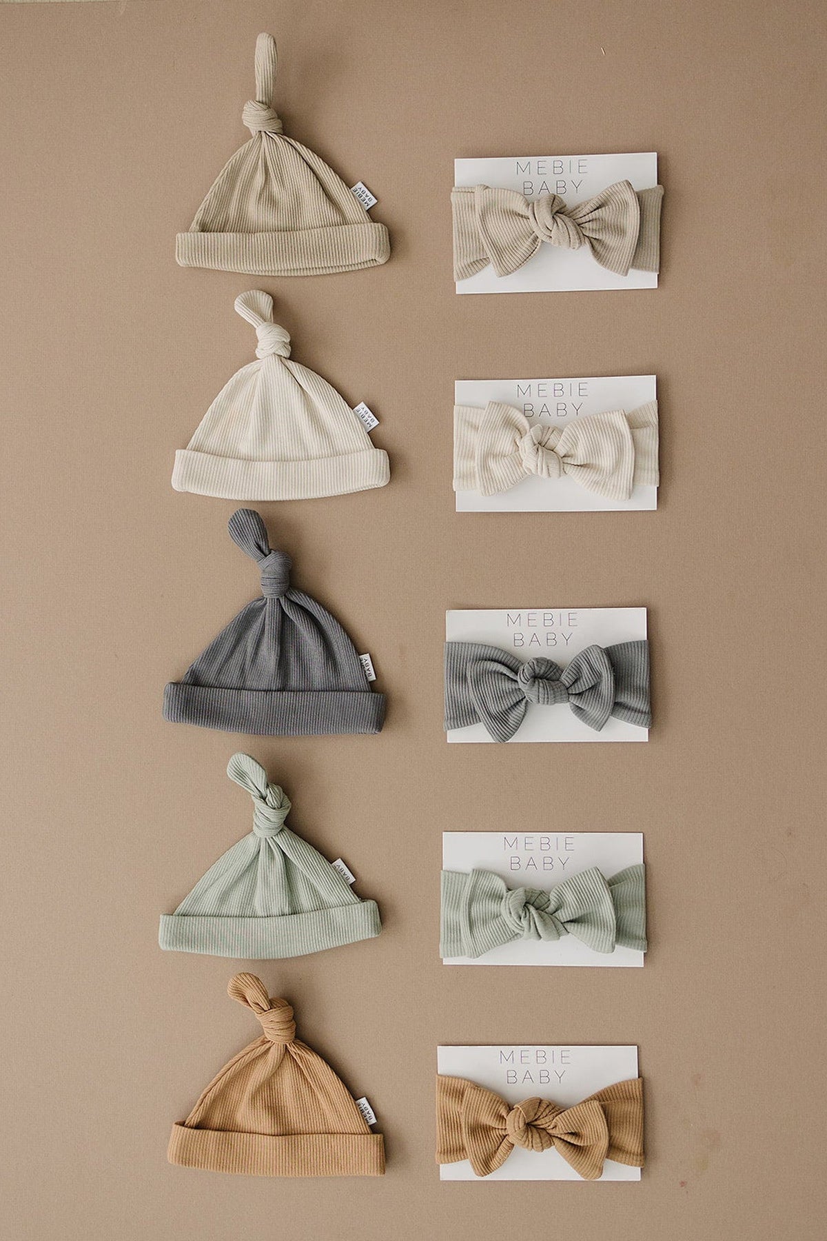 Five ribbed baby hats and matching head wraps in various neutral colors displayed on a brown background.
