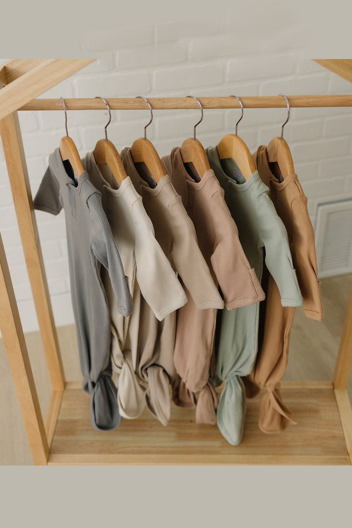 Wooden rack with organic cotton baby knot gowns in neutral colors, hanging on wooden hangers against a brick wall.