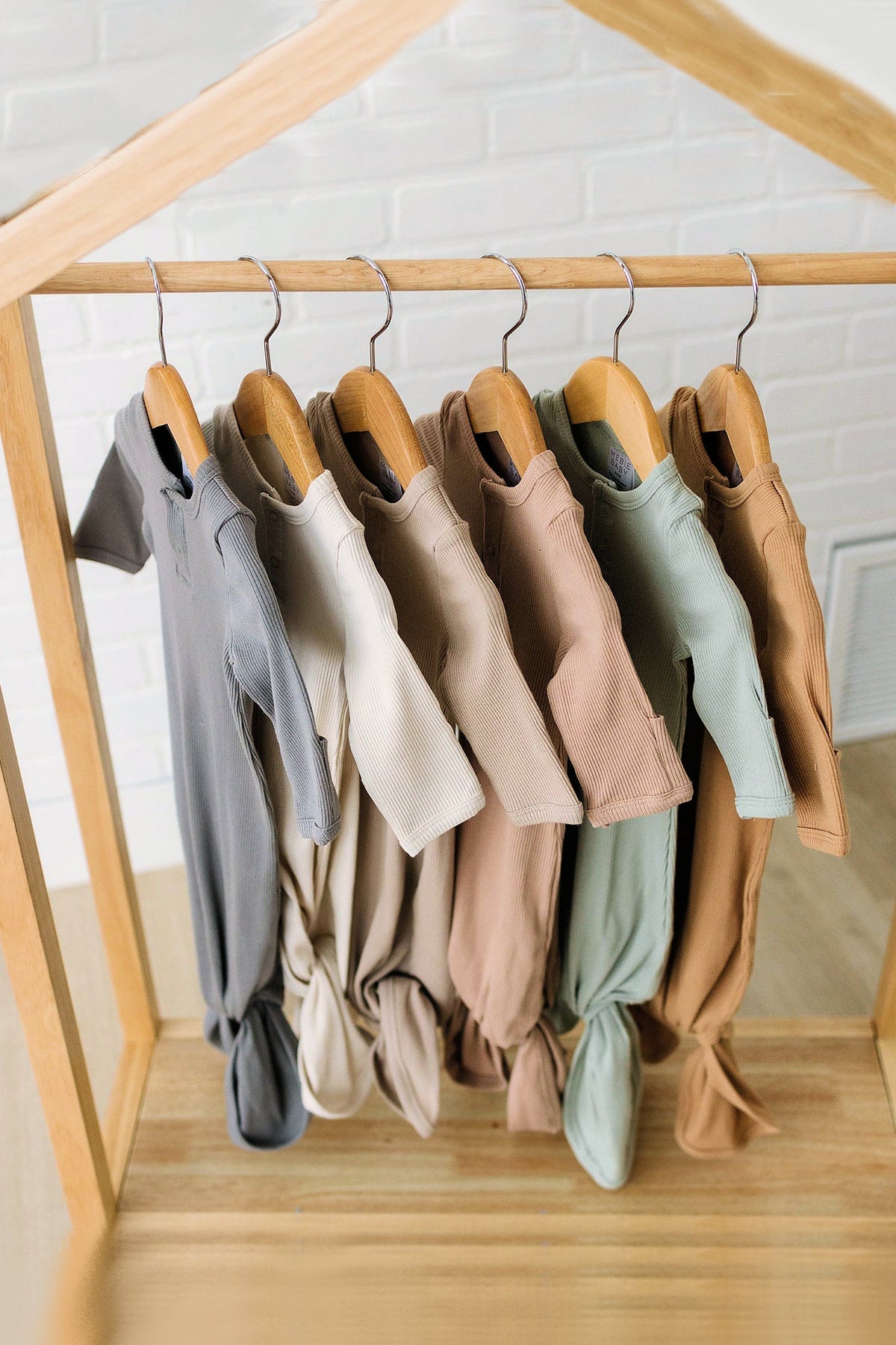 Seven ribbed baby gowns in neutral tones hang on wooden hangers, displayed on a wooden rack.
