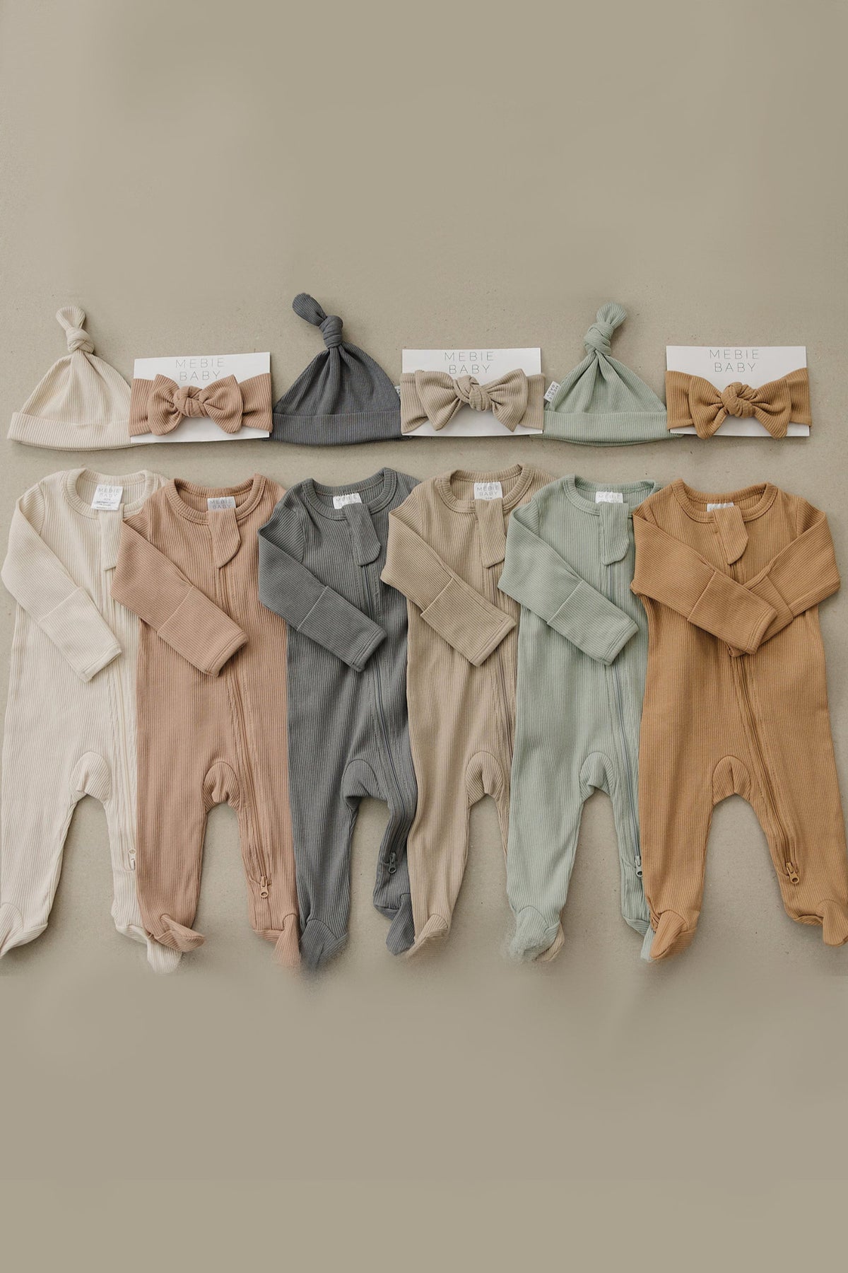 Six ribbed zipper footed baby pajamas in neutral tones with matching knotted hats and bow headbands on display.