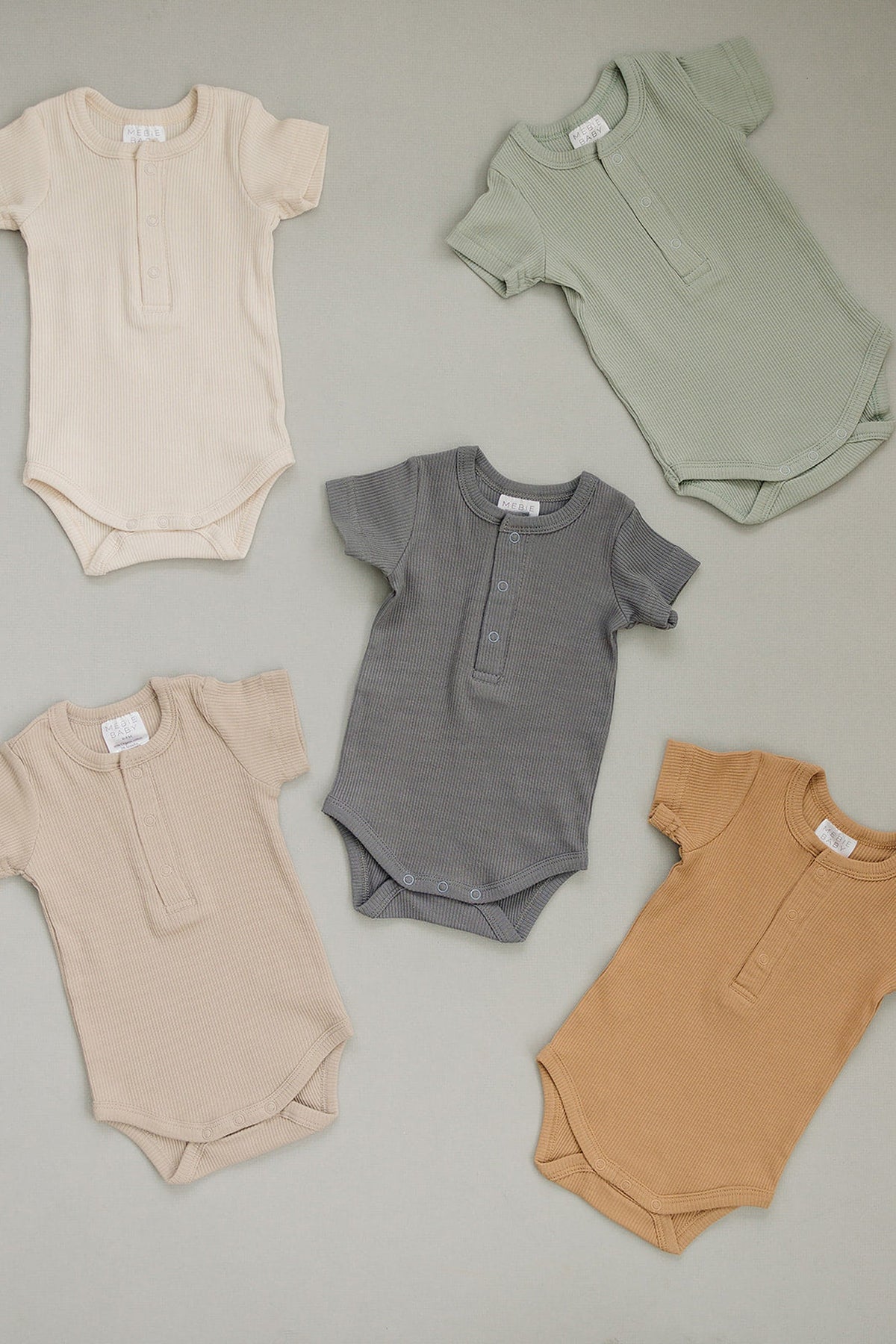 Five ribbed baby bodysuits in earthy tones arranged on a neutral background, featuring short sleeves and snap closures.