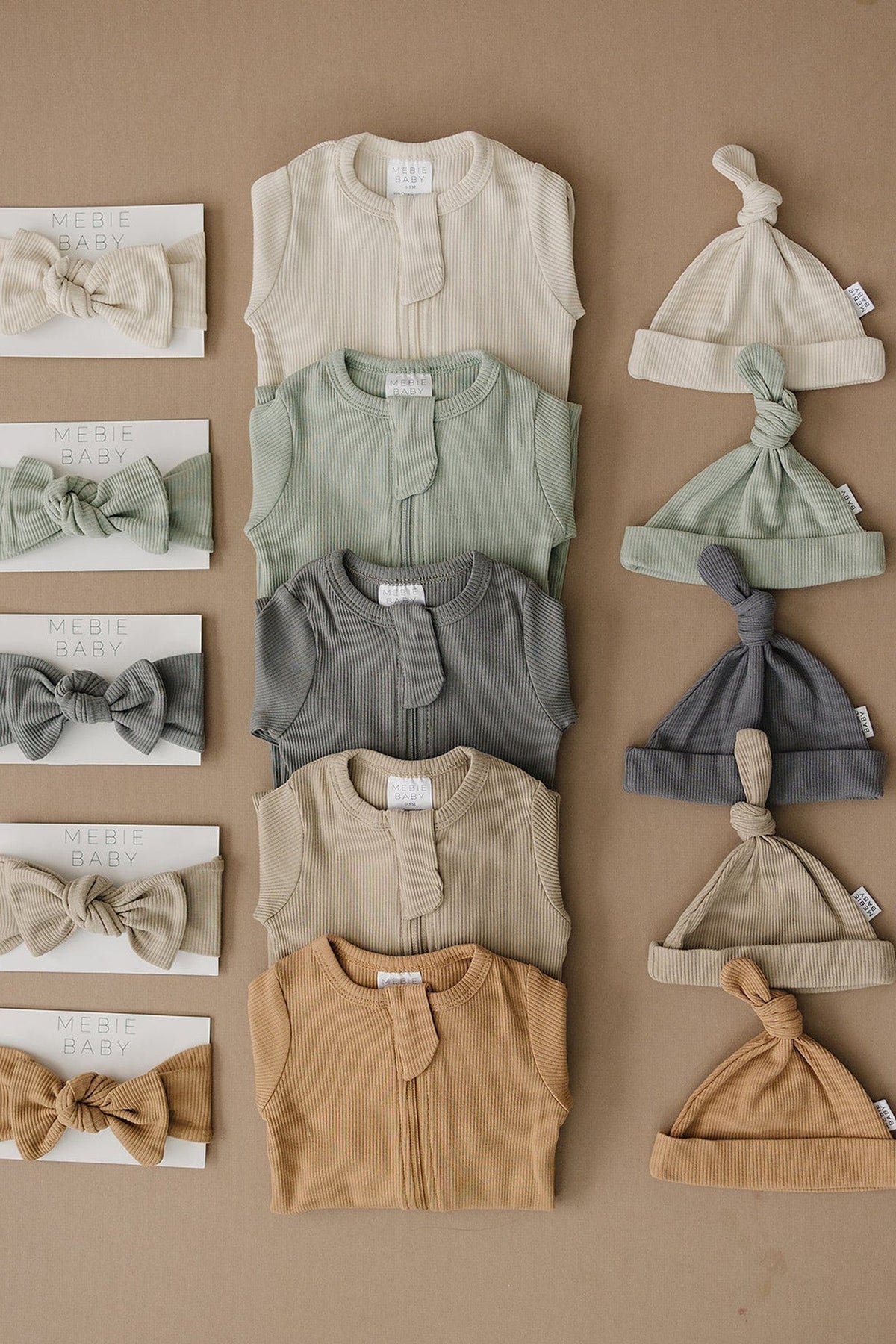 Assorted Mebie Baby ribbed pajamas and matching hats in neutral tones, displayed on a brown background.