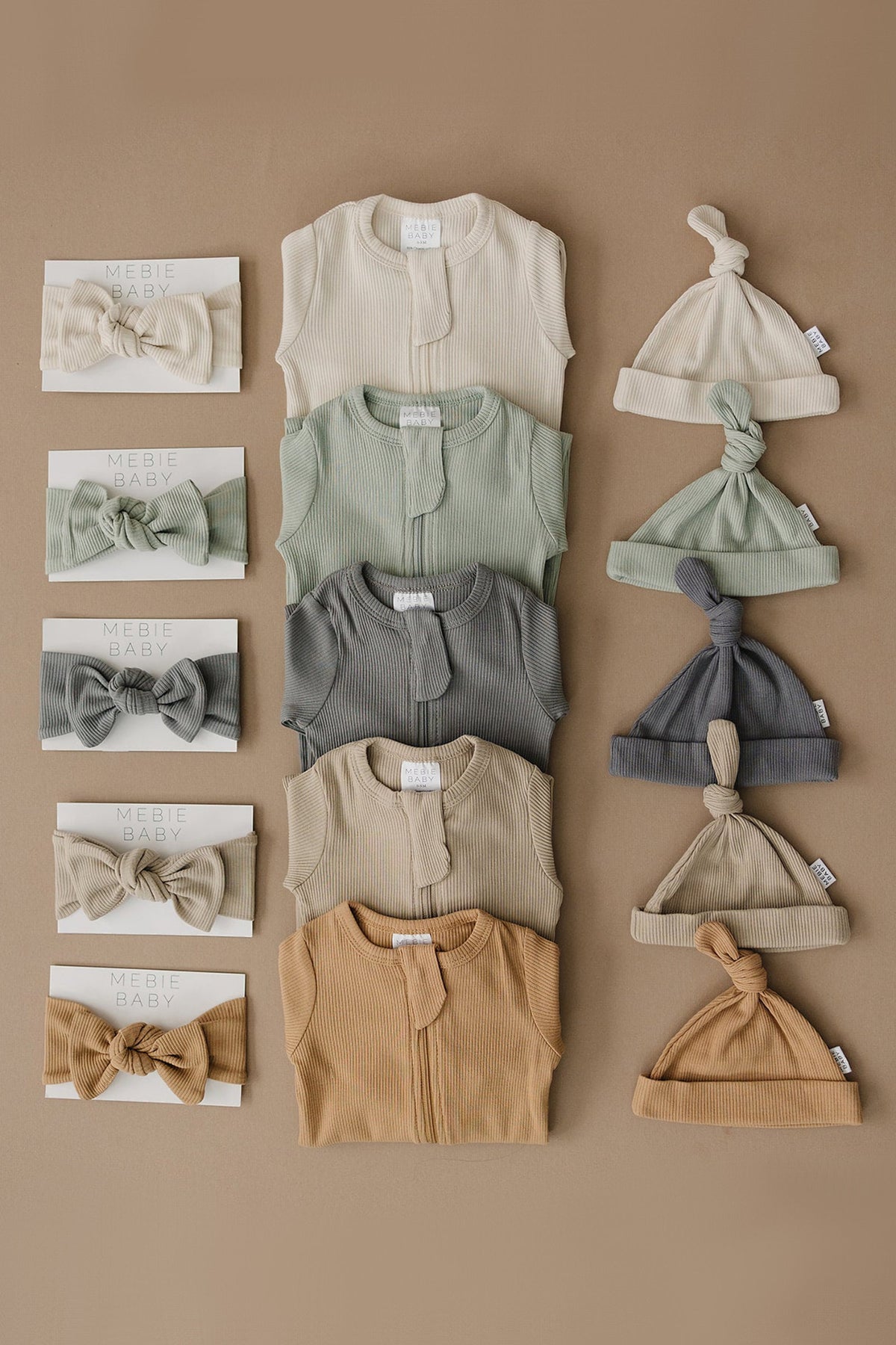 Assorted Mebie Baby ribbed clothing sets in neutral colors, including onesies, hats, and headbands on a brown background.