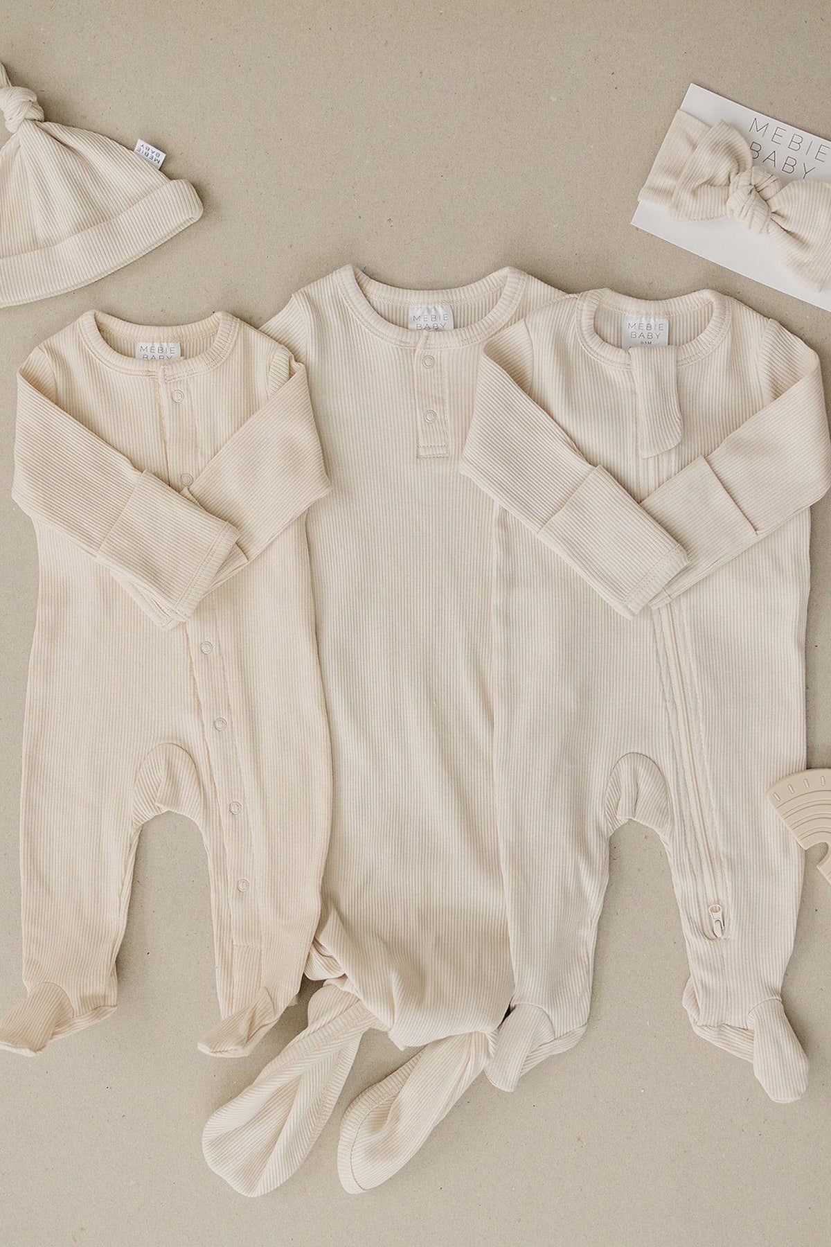 Three vanilla ribbed baby pajamas with footed bottoms and snaps, accompanied by a matching hat and bow.