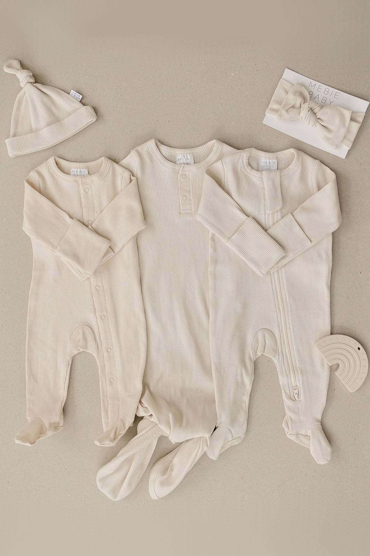 Three vanilla ribbed baby gowns with matching hat and headband, made from soft organic cotton, displayed on beige background.