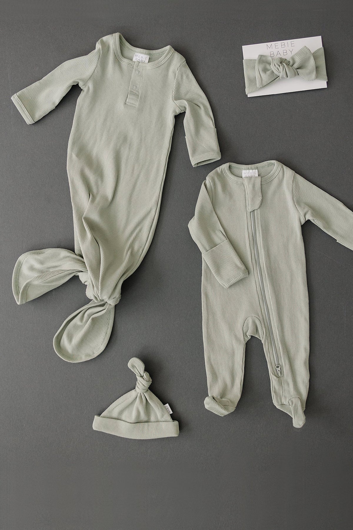 Sage ribbed baby clothing set includes a knotted gown, footed pajamas, hat, and headband on a dark background.