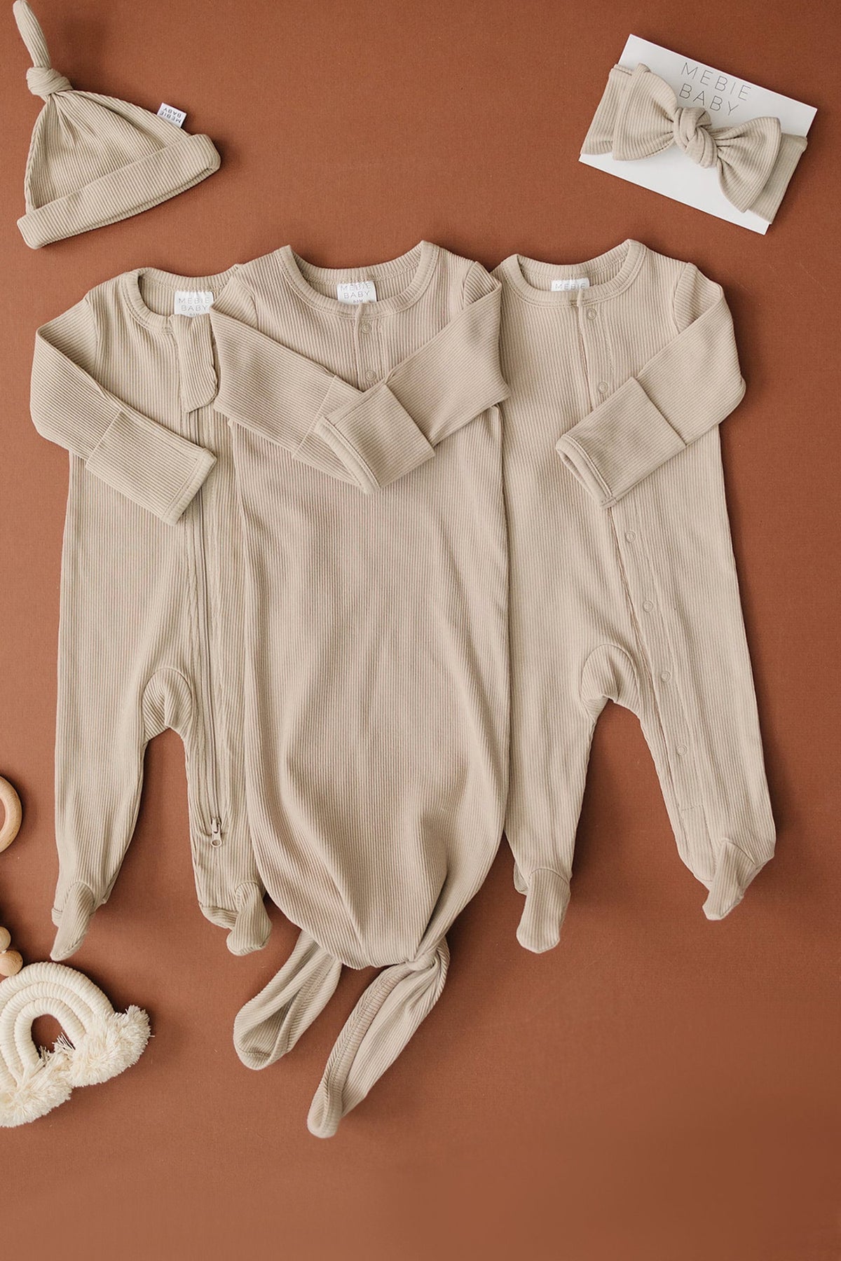 Three oatmeal-colored ribbed baby pajamas with footed bottoms, a matching hat, and headband on a brown surface.