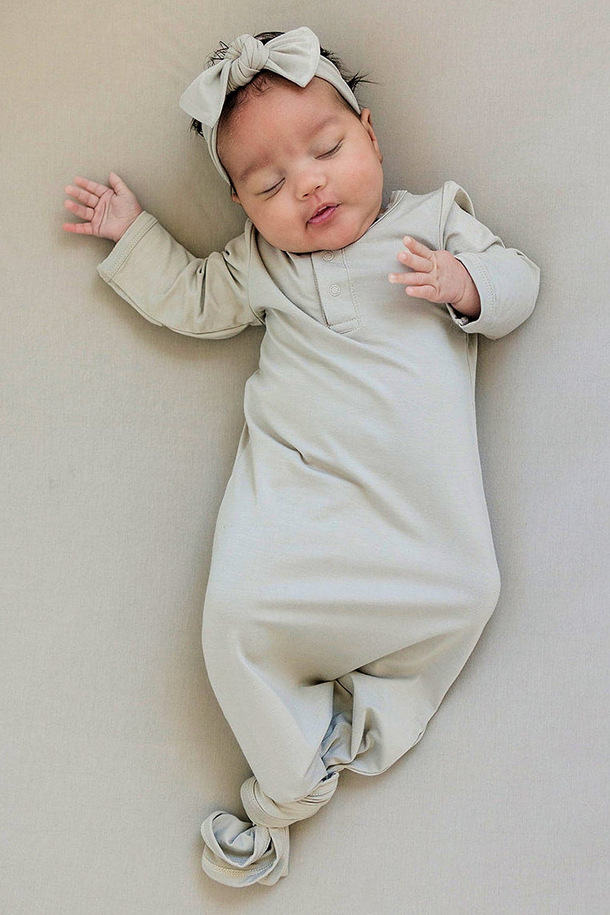A baby wearing an oatmeal-colored knotted gown and matching head wrap sleeps peacefully on a neutral background.