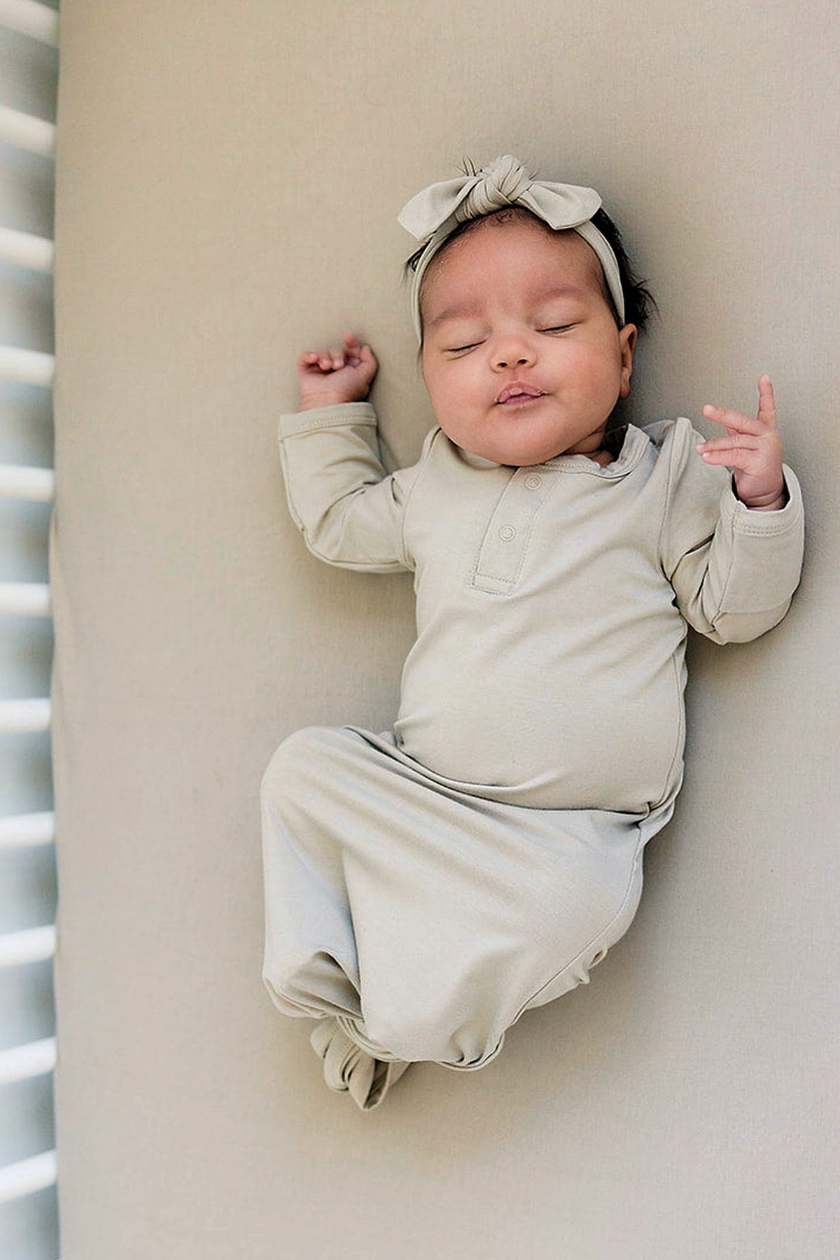 A baby wearing an oatmeal-colored bamboo knot gown and matching headband lies peacefully on a soft surface.