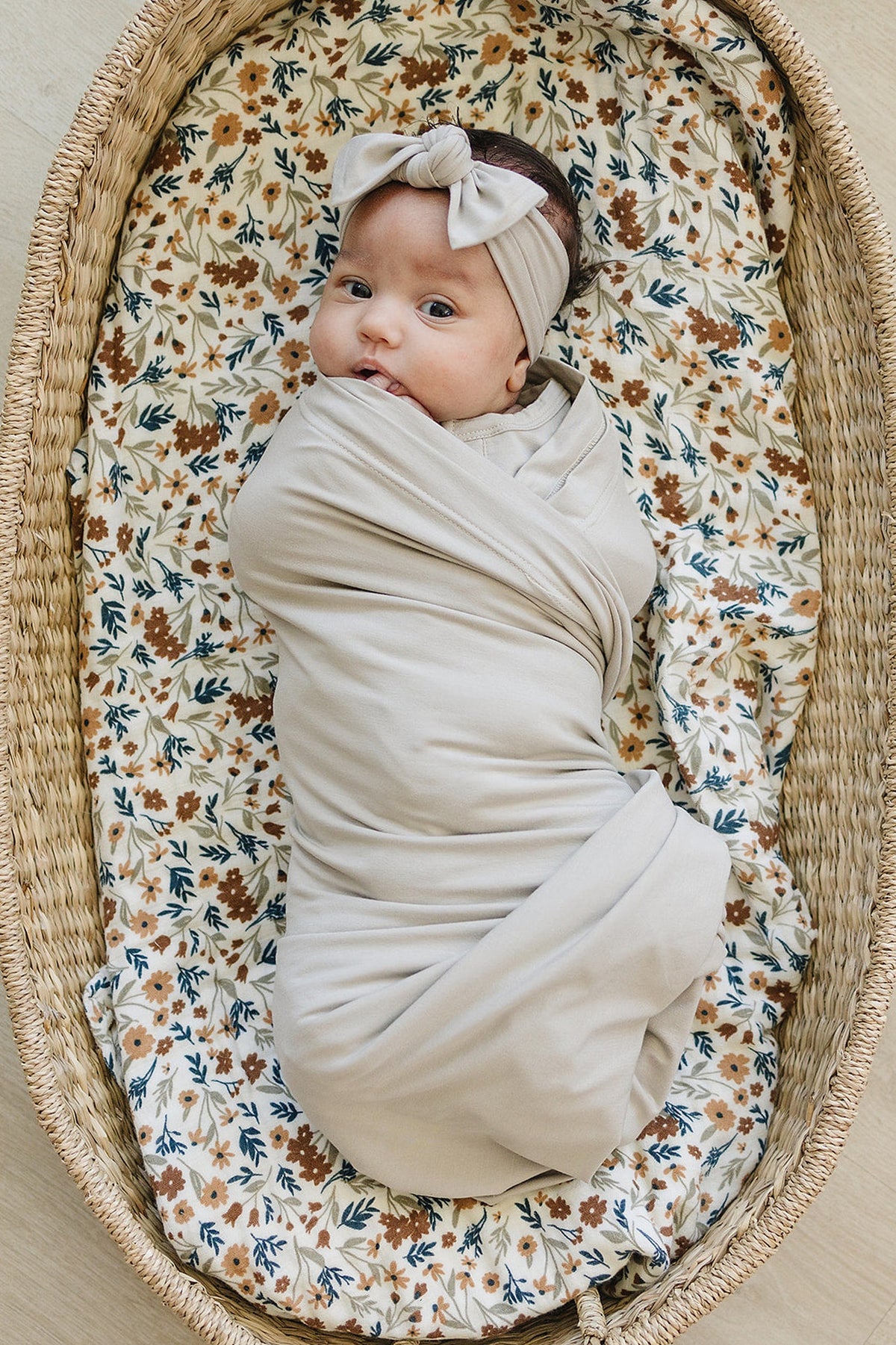 A baby wrapped in a beige swaddle blanket, lying in a wicker basket with a floral-patterned liner.