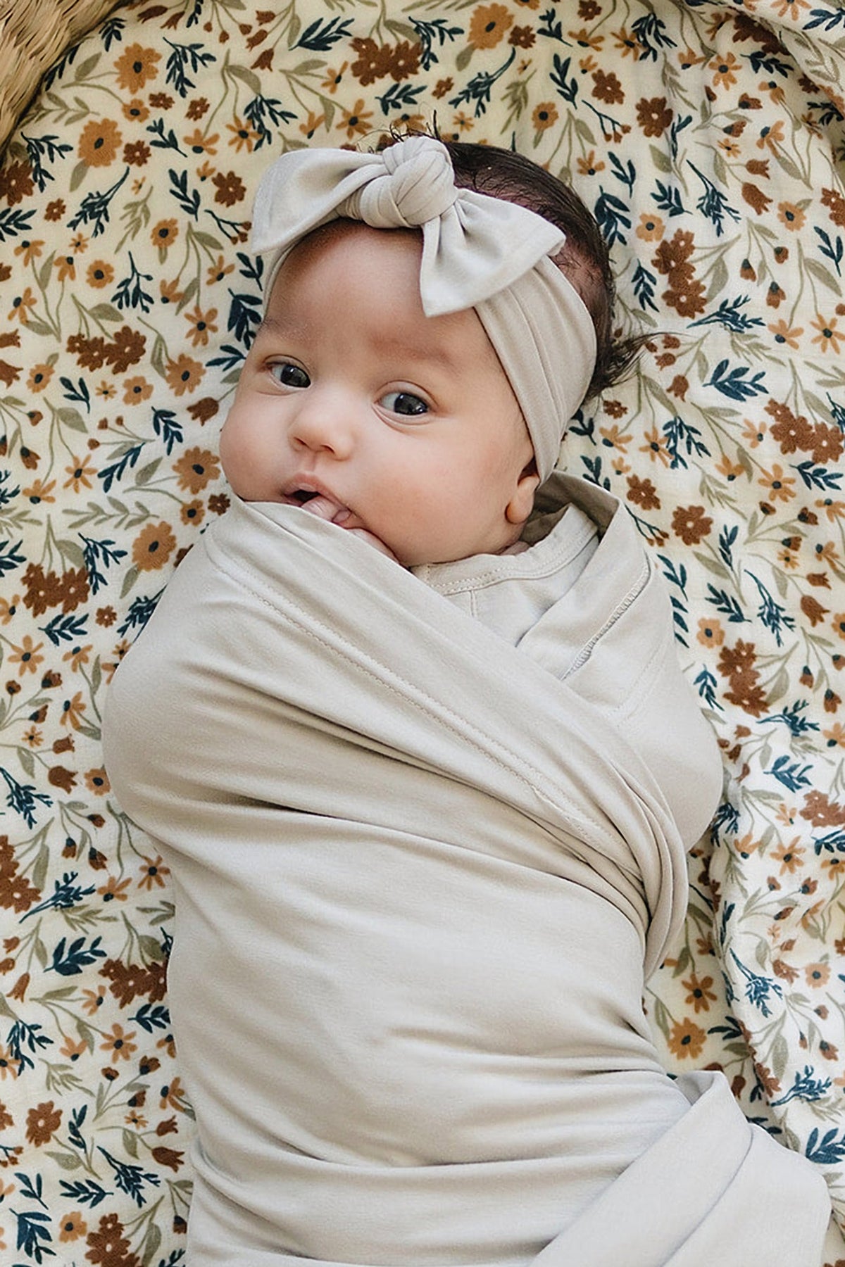 A baby wrapped in an oatmeal-colored swaddle with a matching head wrap, lying on a floral-patterned sheet.