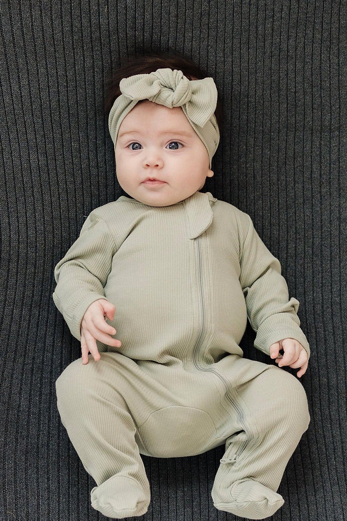 A baby in sage ribbed pajamas and matching headband lies on a dark ribbed fabric background.