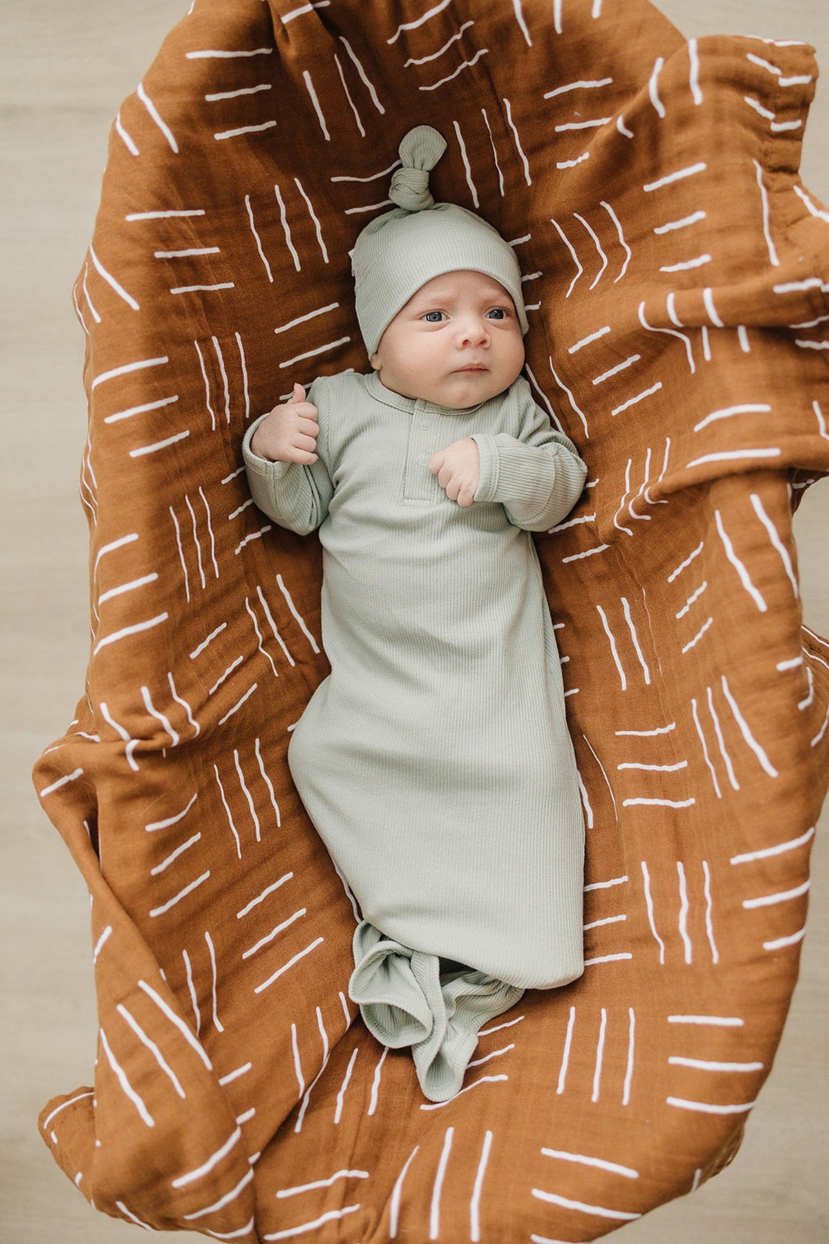A newborn wearing a sage ribbed knot hat and gown lies on a brown patterned blanket.