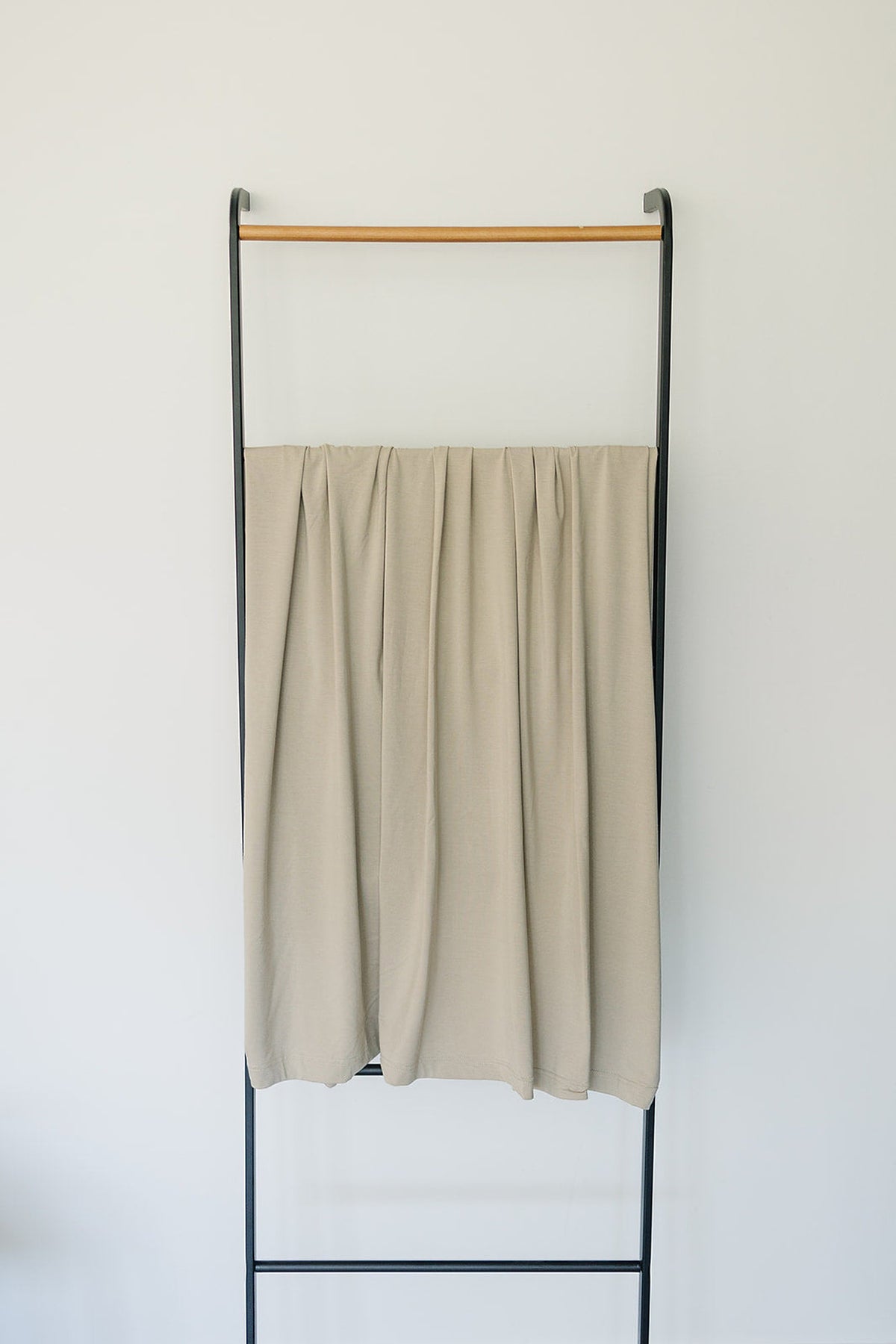 Beige swaddle blanket made from bamboo fabric draped over a black metal and wooden rack against a white wall.