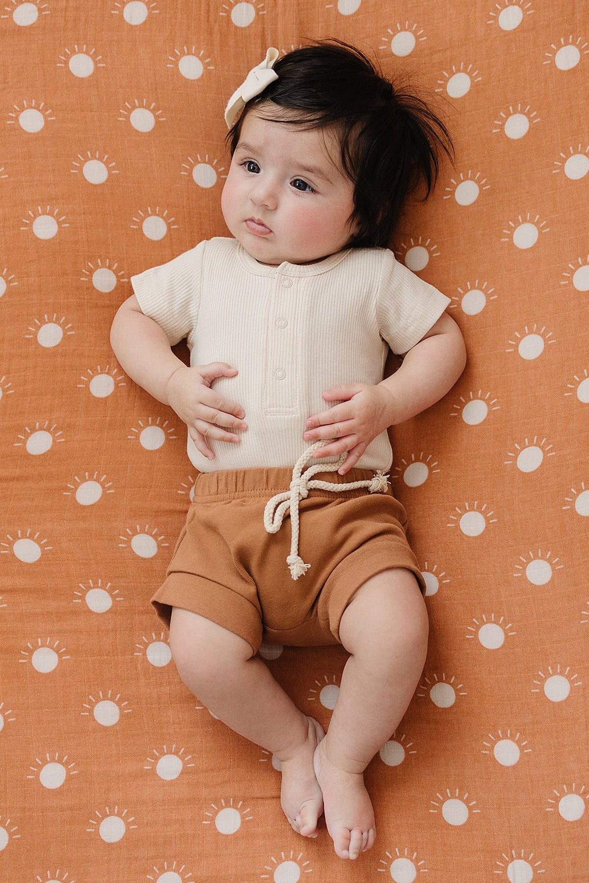 A baby in a cream ribbed bodysuit and brown shorts lies on an orange sun-patterned blanket.