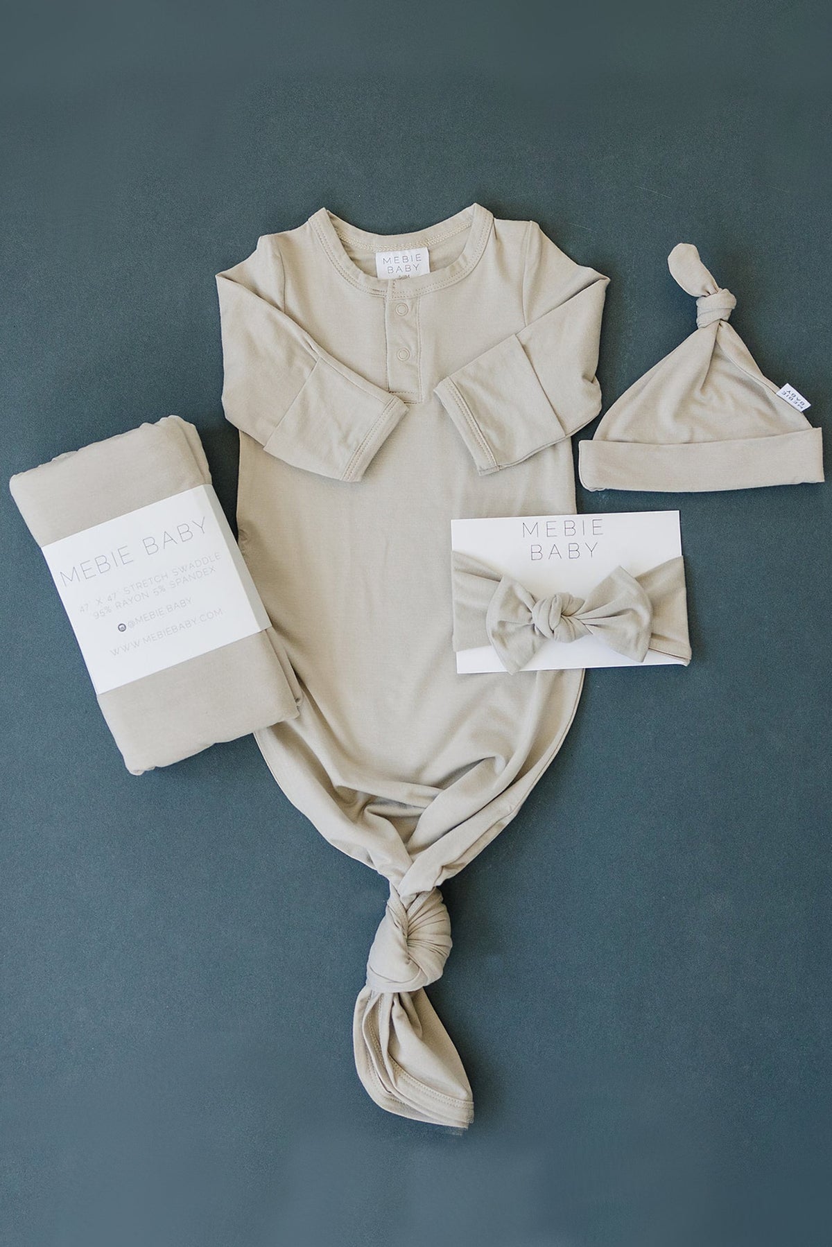 Beige baby outfit set with a knotted gown, hat, swaddle, and bow headband on a dark background.