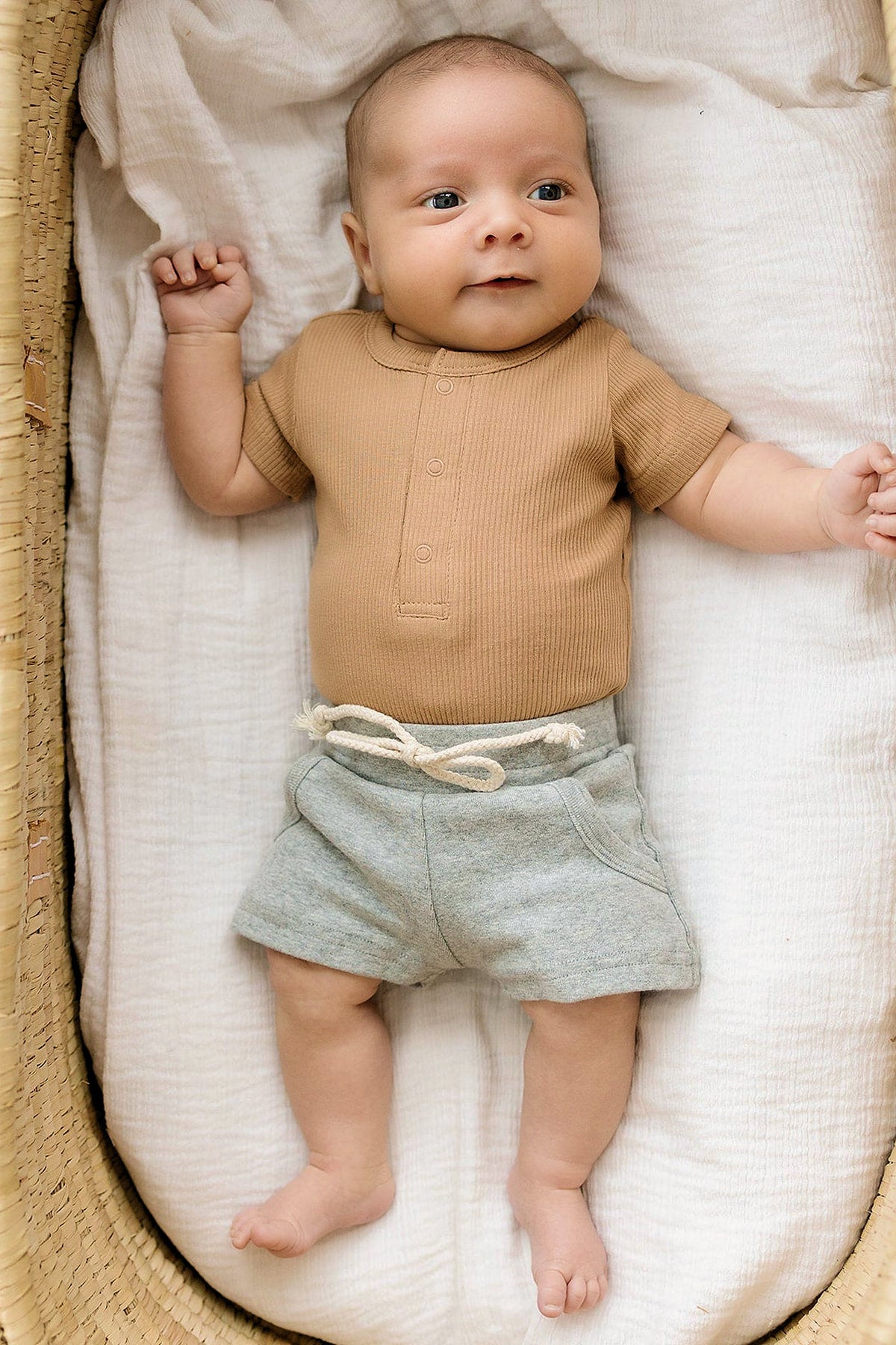 Baby in a tan ribbed bodysuit and gray shorts lies on a white blanket in a woven basket.