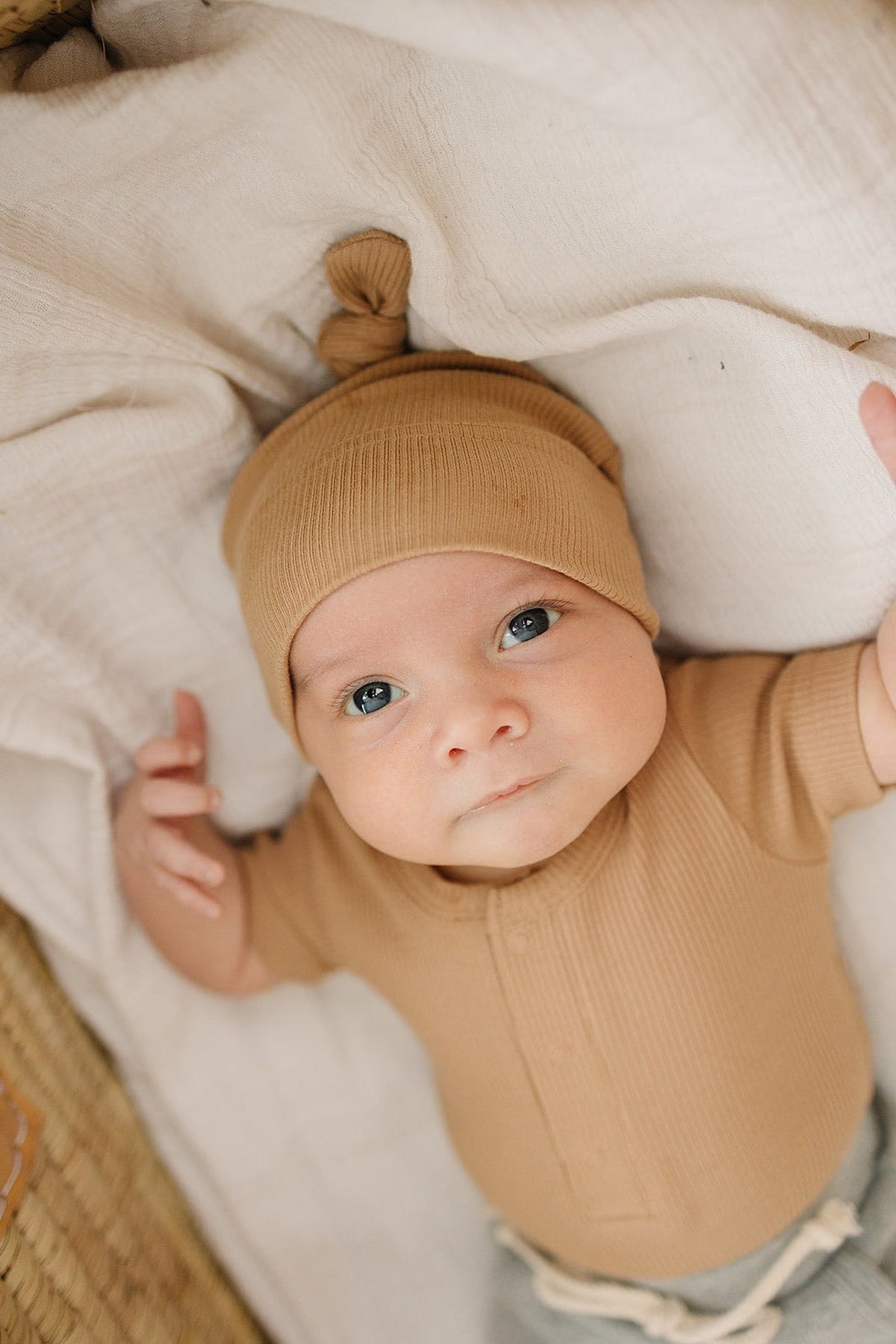 A baby wearing a brown ribbed knot hat and matching outfit lies on a soft white blanket.