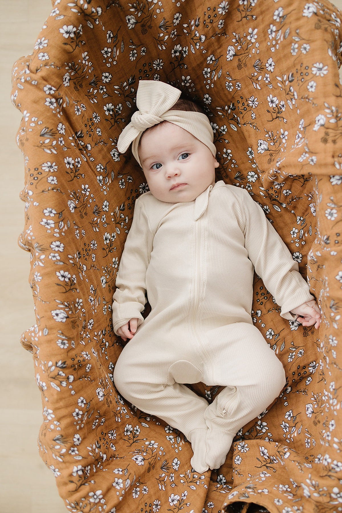 A baby in a cream ribbed footed pajama lies on a floral-patterned brown blanket, wearing a matching headband.