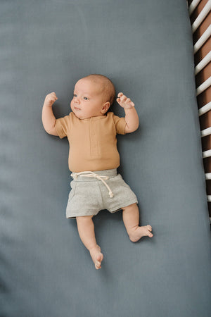 A baby in a beige shirt and gray shorts lies on a dusty blue crib sheet.