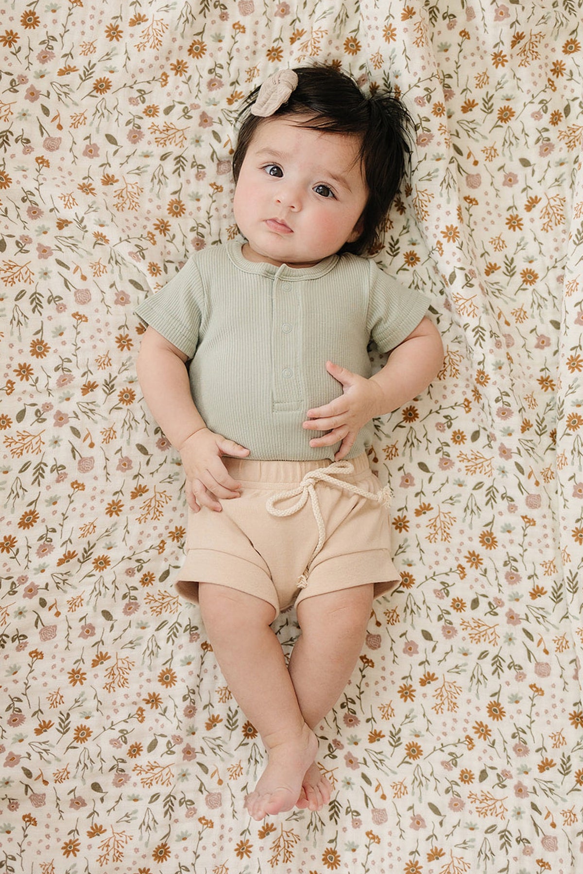A baby wearing a sage ribbed bodysuit and beige shorts lies on a floral-patterned blanket.