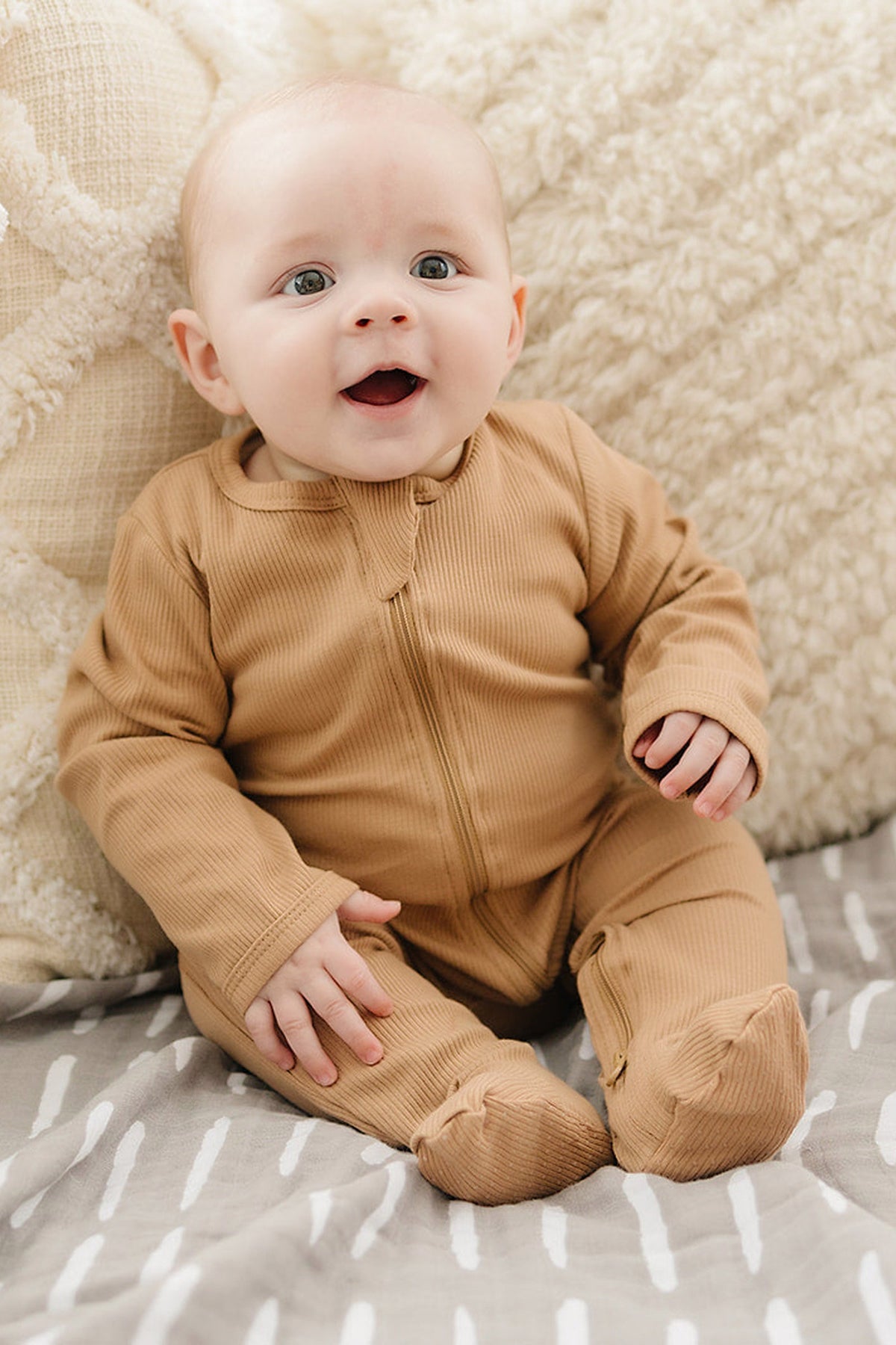 A baby wearing tan ribbed zipper footed pajamas sits on a cozy blanket, smiling joyfully.