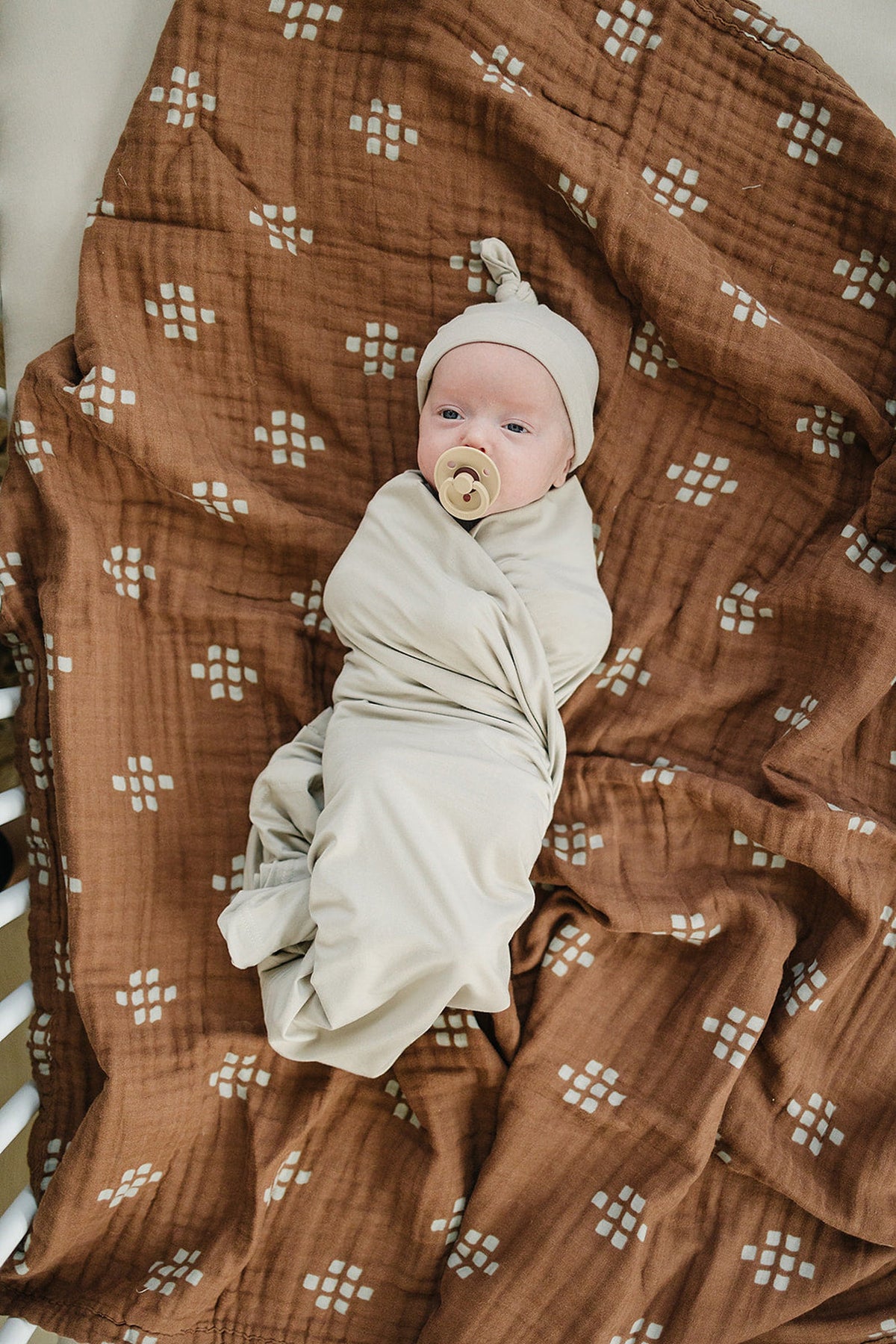 Baby wrapped in a beige bamboo swaddle, wearing a matching hat, lying on a patterned brown blanket.