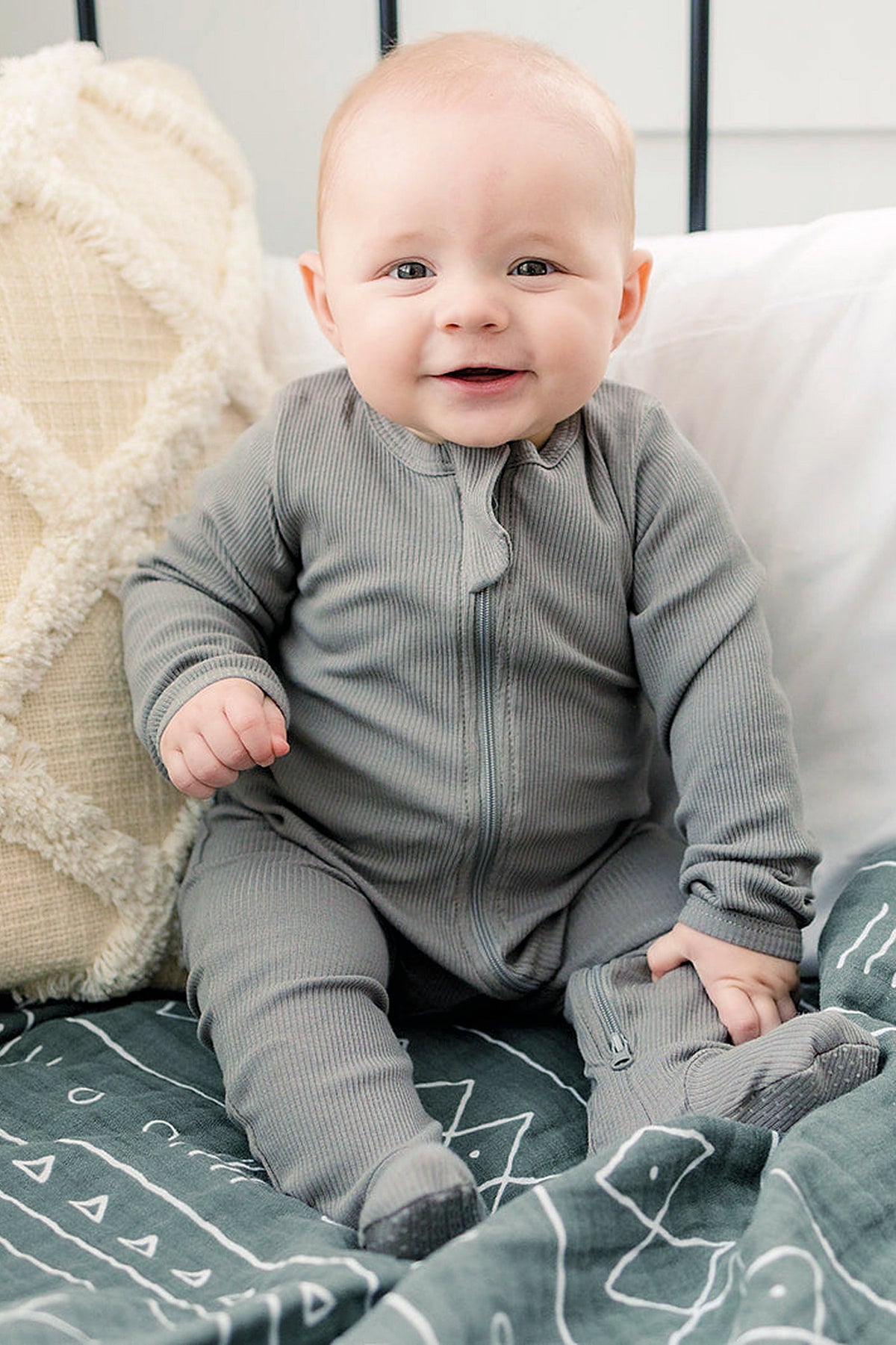 A baby in a grey ribbed zipper footed pajama sits on a patterned blanket, smiling.