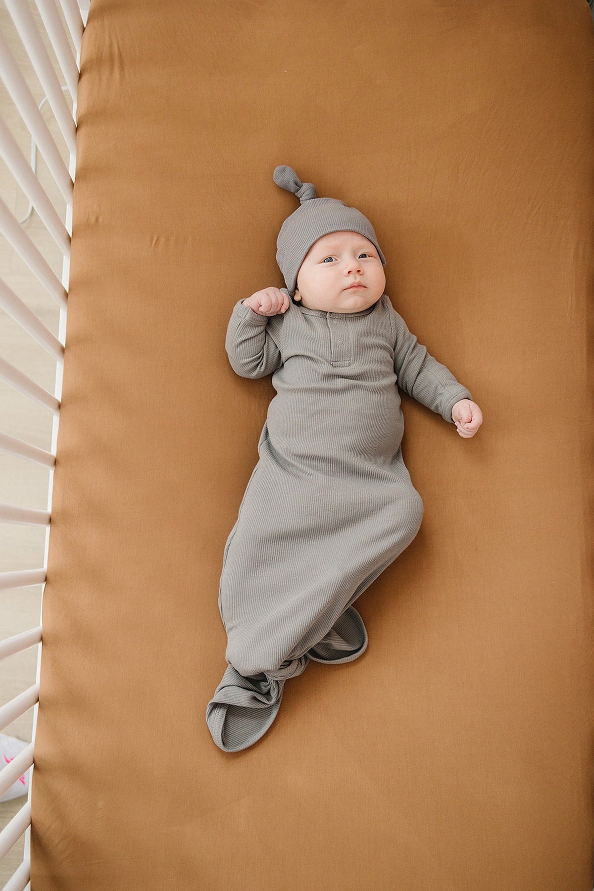 A baby in a gray outfit lies on a mustard crib sheet in a white crib.