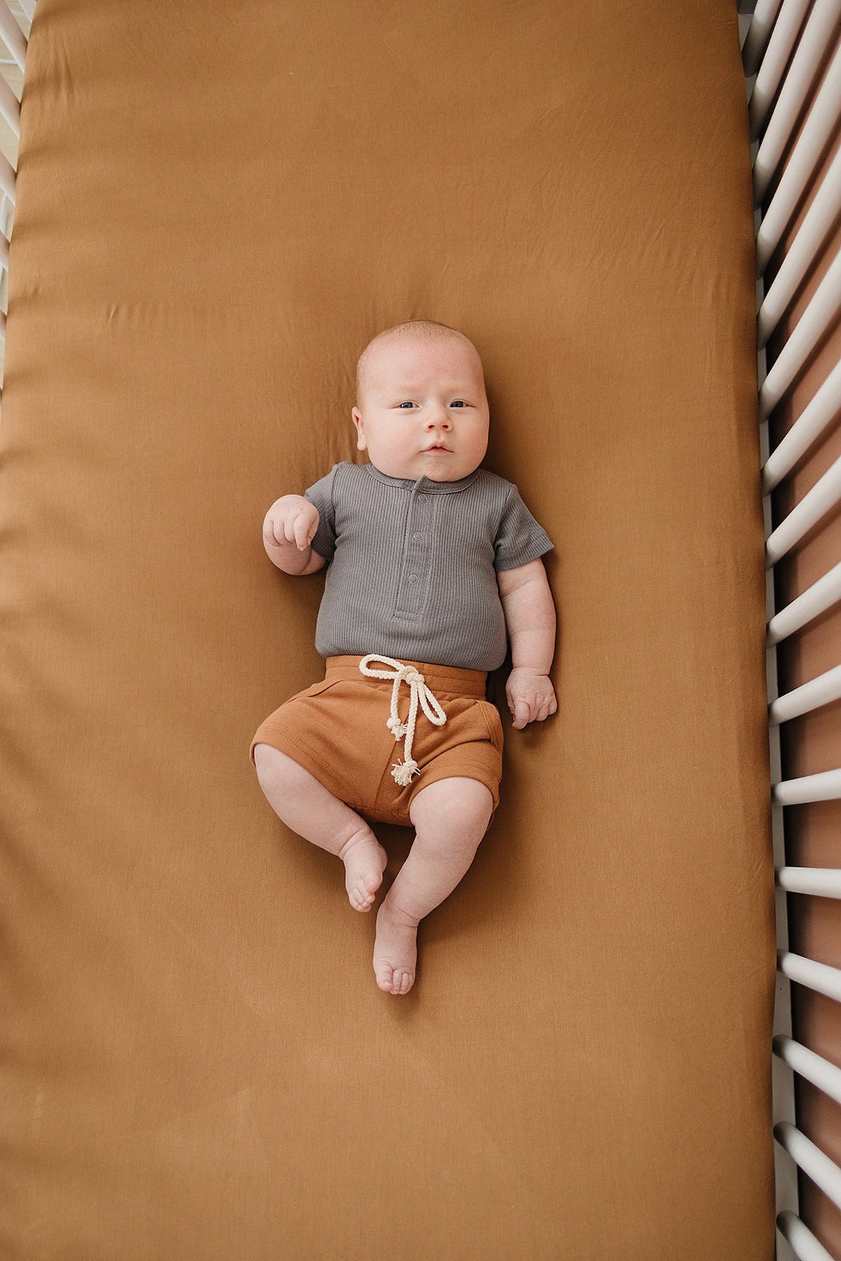 A baby in a grey shirt and mustard shorts lies on a mustard-colored crib sheet inside a crib.