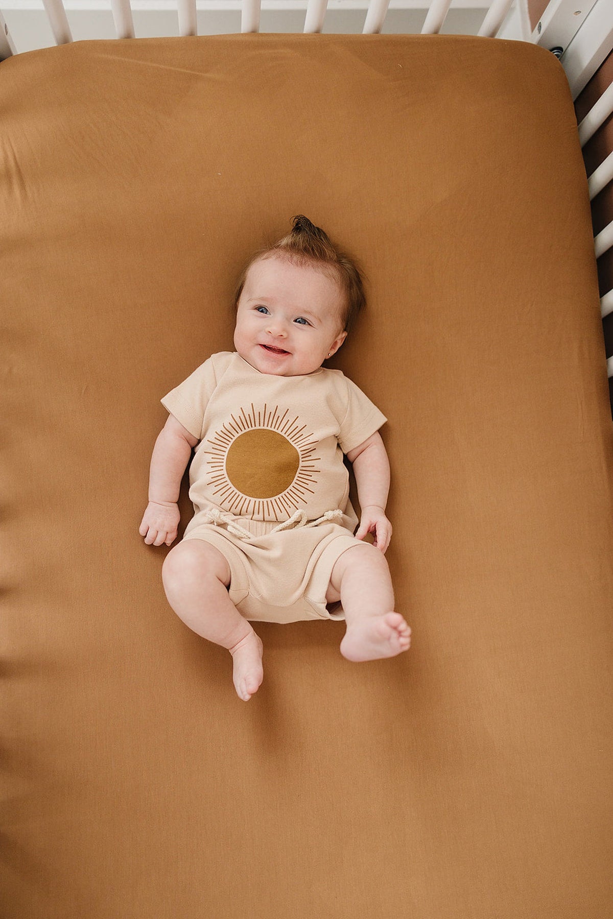 A smiling baby lies on a mustard-colored crib sheet, wearing a beige outfit with a sun design.
