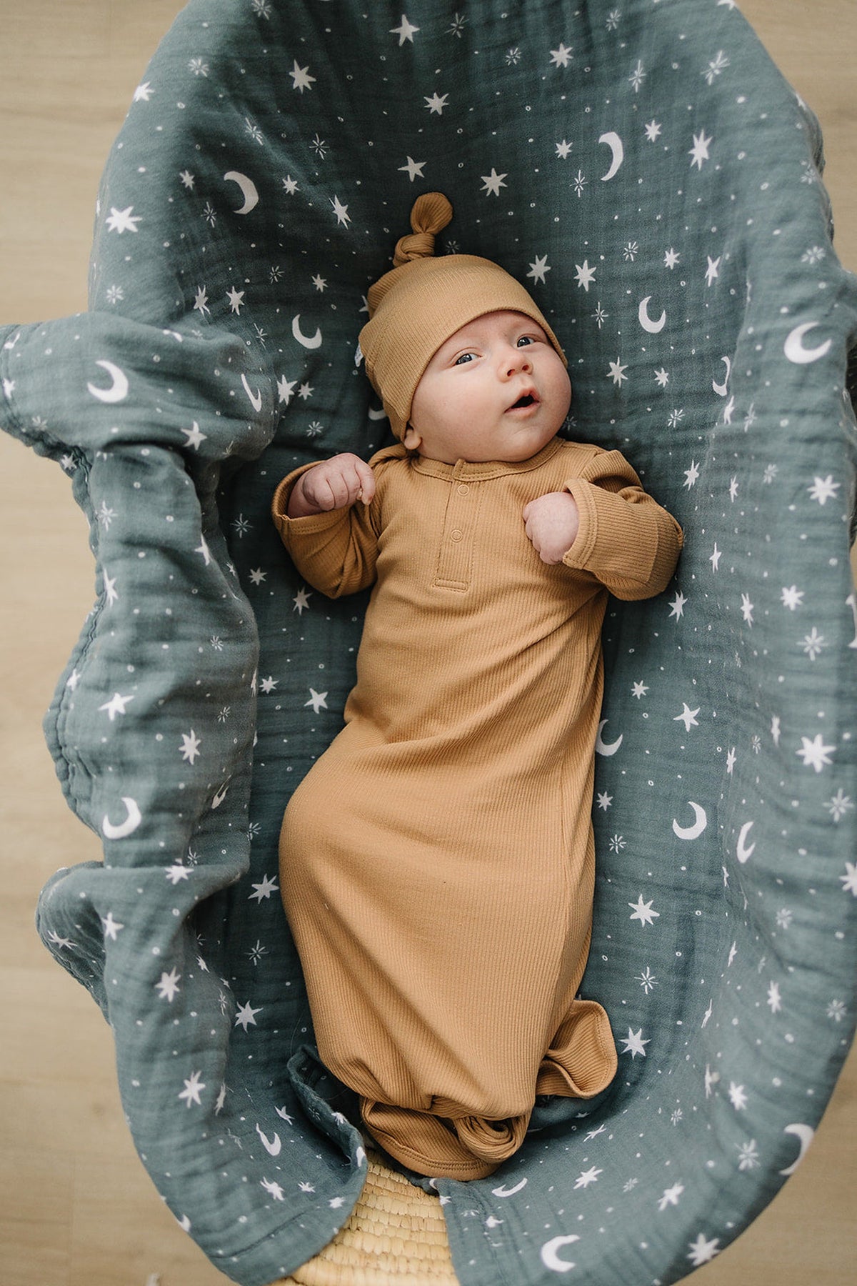 A baby in a tan ribbed gown and knot hat lies on a celestial-patterned green blanket.