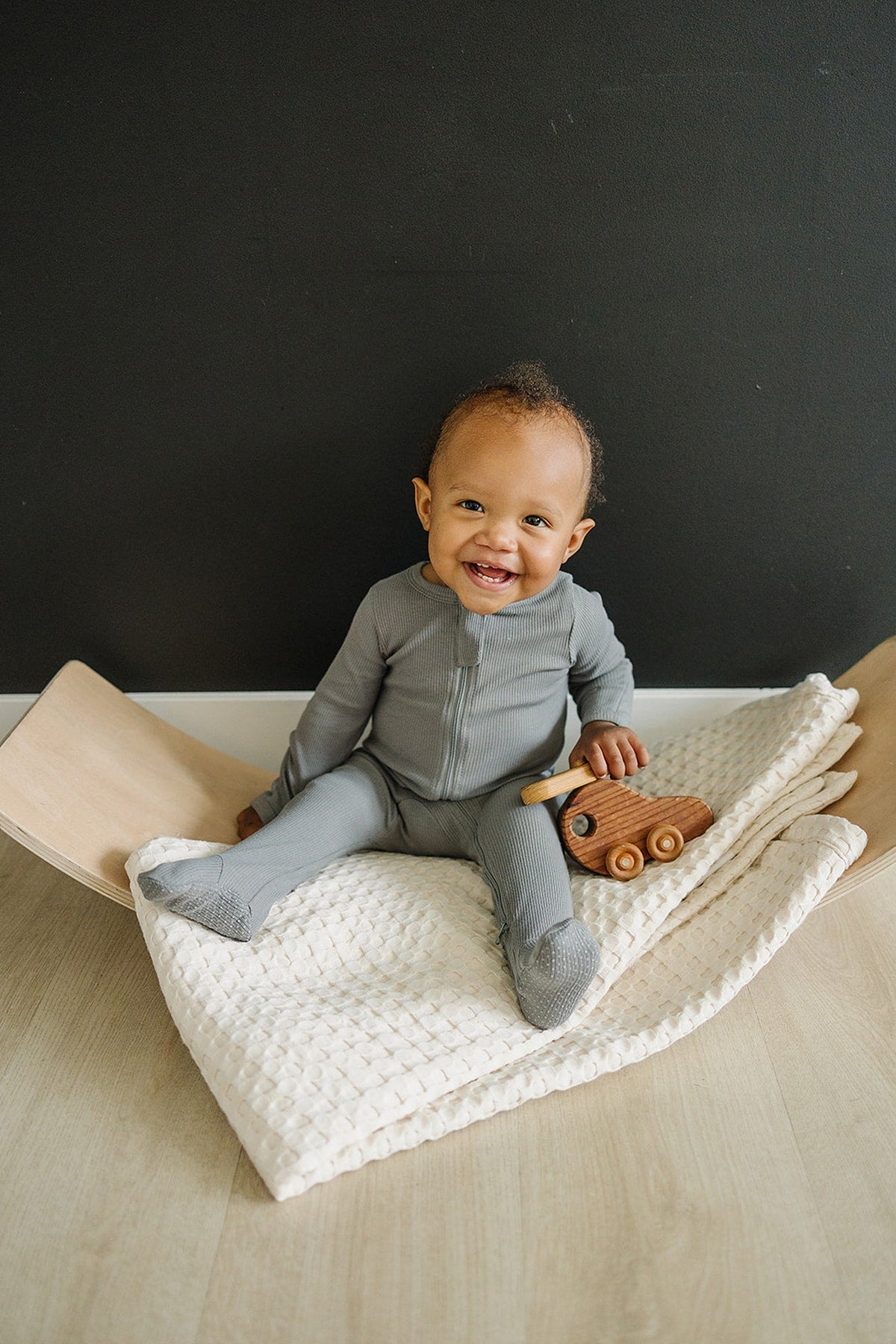 A baby wearing grey ribbed zipper footed pajamas sits on a cream-textured blanket, holding a wooden toy car.
