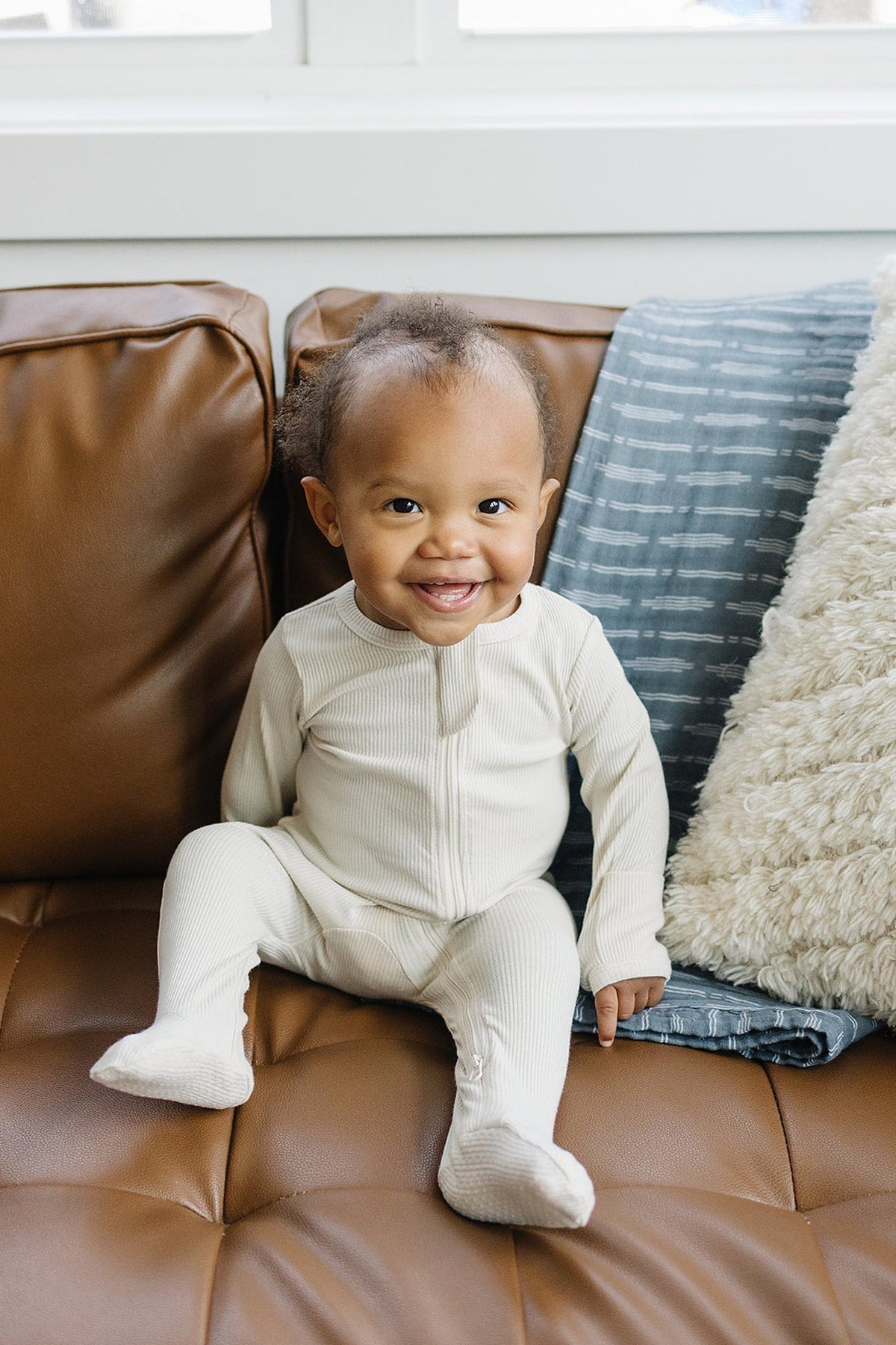 A baby smiles while sitting on a brown leather couch, wearing a cream-colored ribbed footed pajama with a zipper.