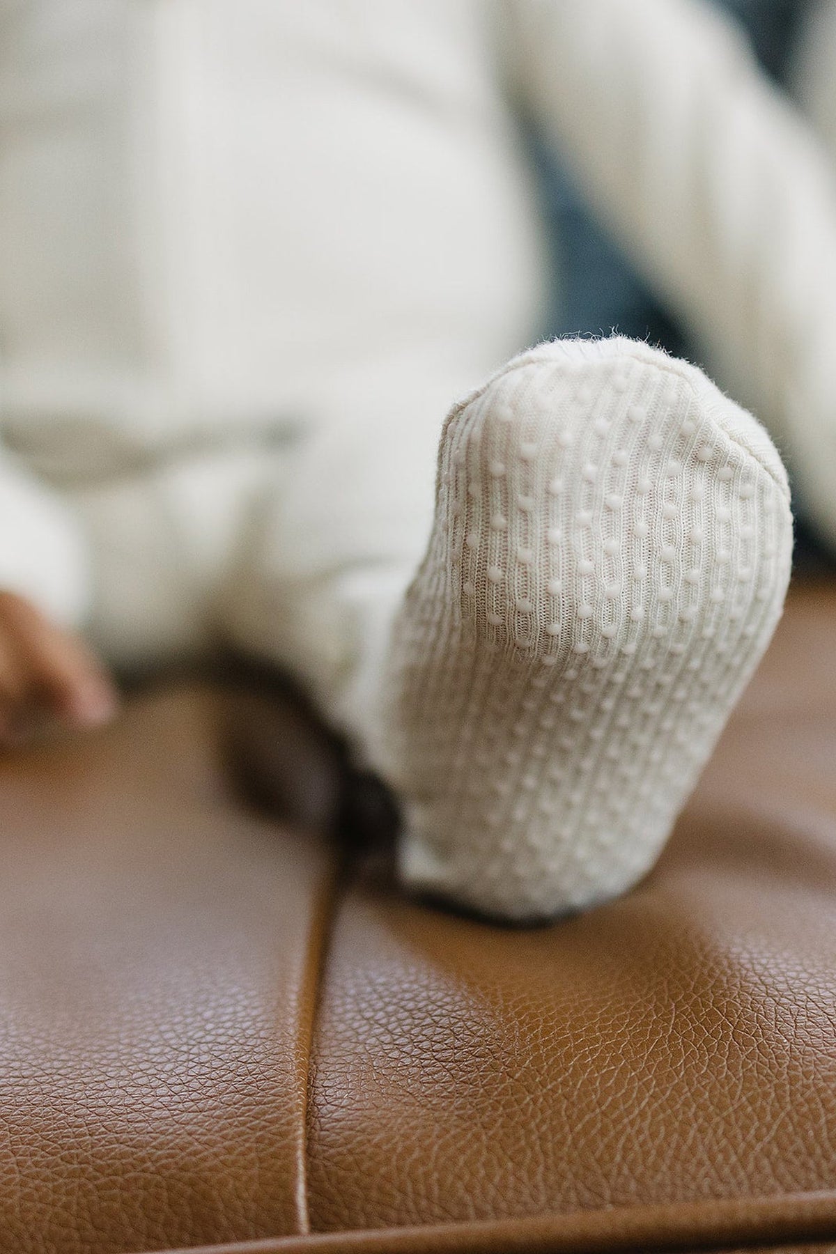 A close-up of a babys foot, wearing a soft, ribbed, cream-colored sock, resting on a brown leather surface.