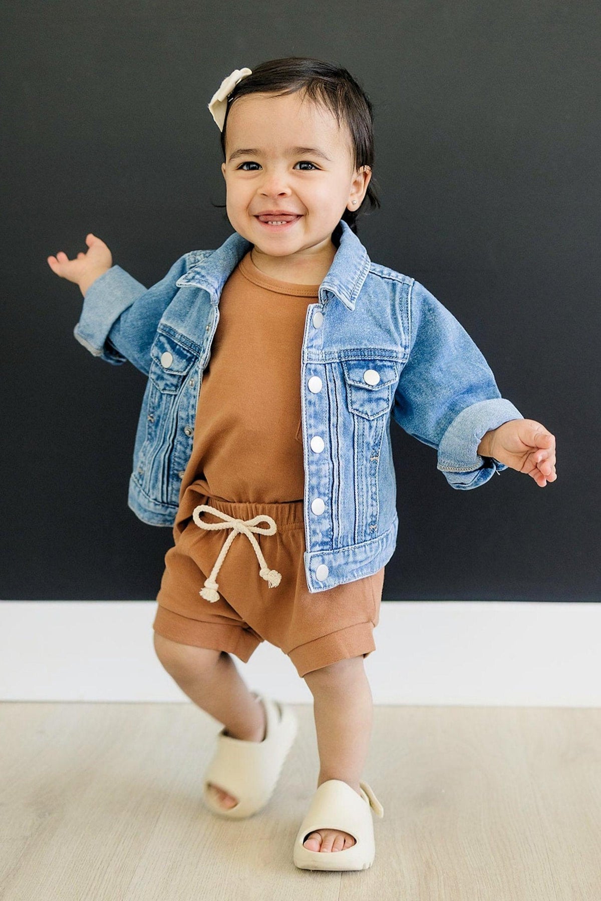 A smiling toddler wearing a light blue denim jacket and brown outfit stands on a light wood floor.