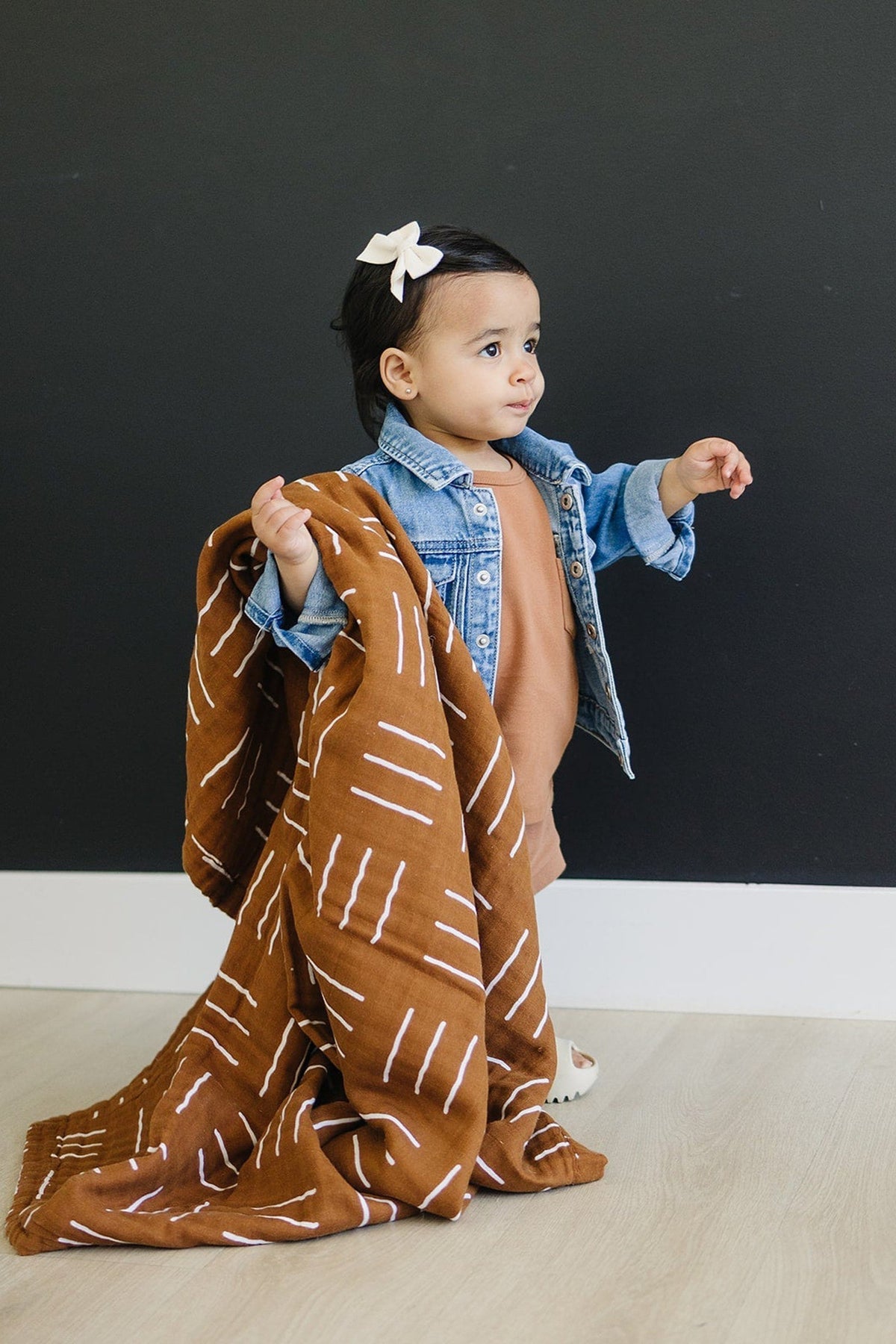 A toddler in a denim jacket holds a mustard mudcloth muslin quilt, standing against a dark wall.