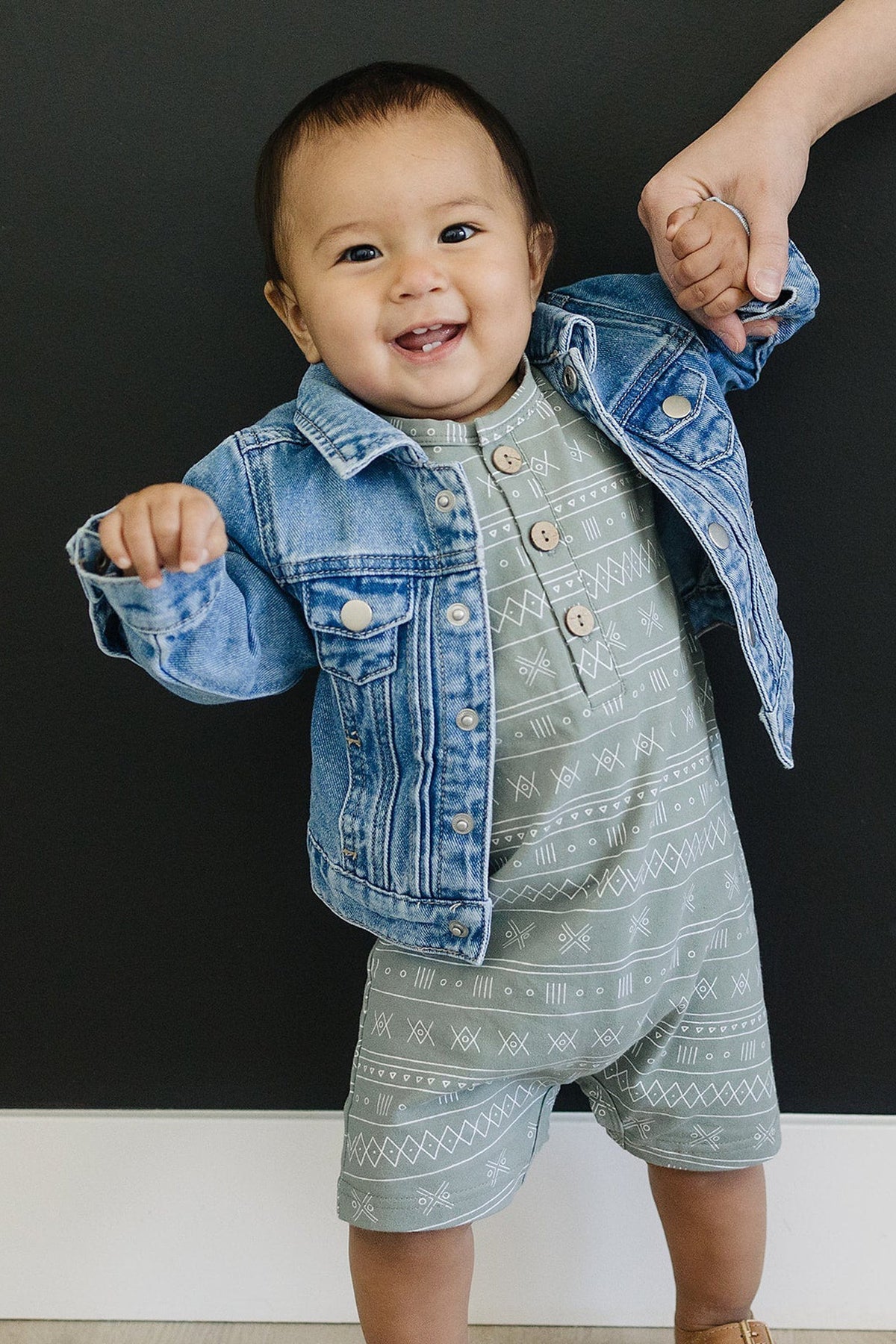 A smiling baby wearing a blue denim jacket and patterned romper, holding an adults hand against a dark background.